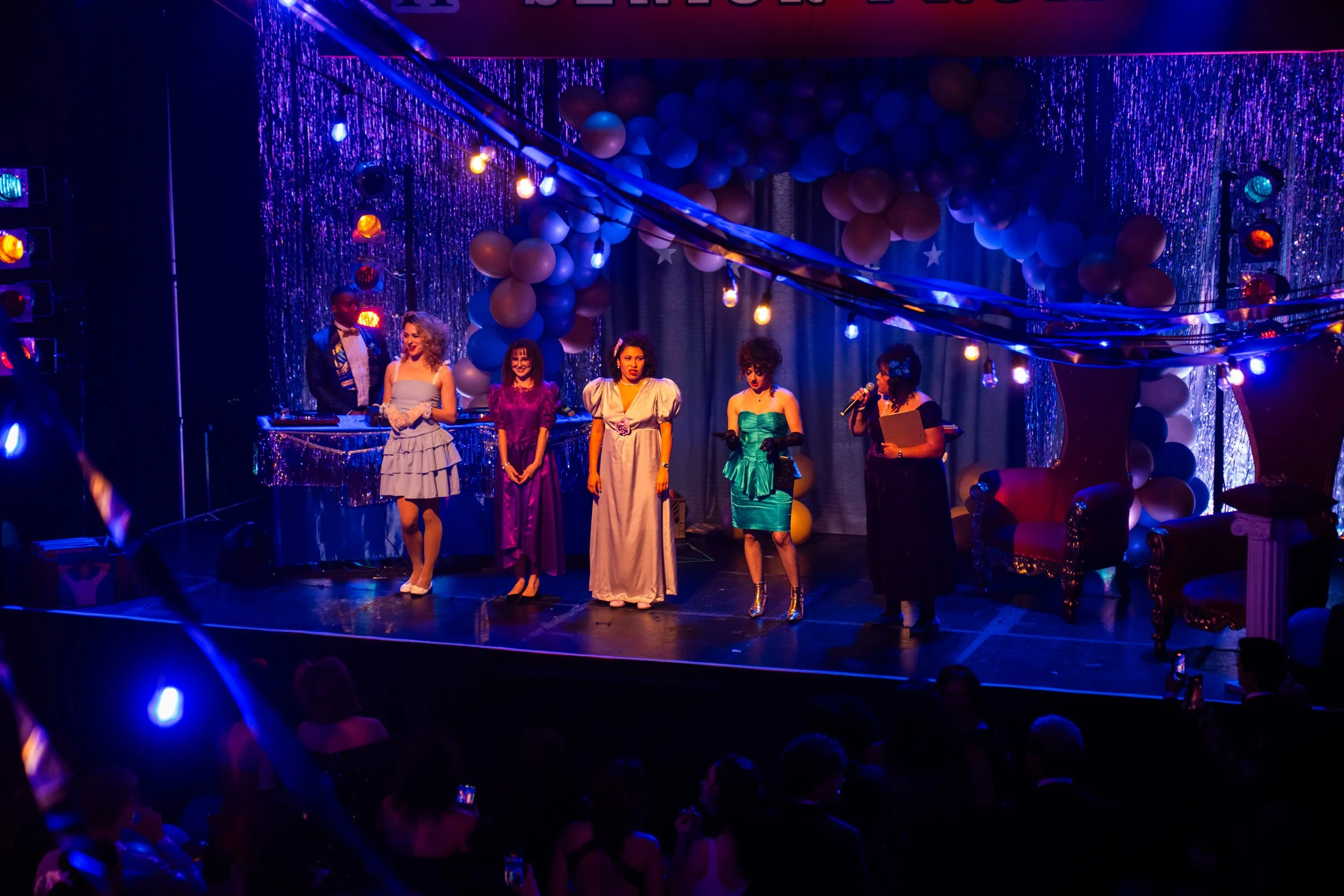 Five women on stage at a theatrical event, dressed in colorful vintage-style dresses, with balloons and streamers decorating the background, and an audience watching in dim lighting.
