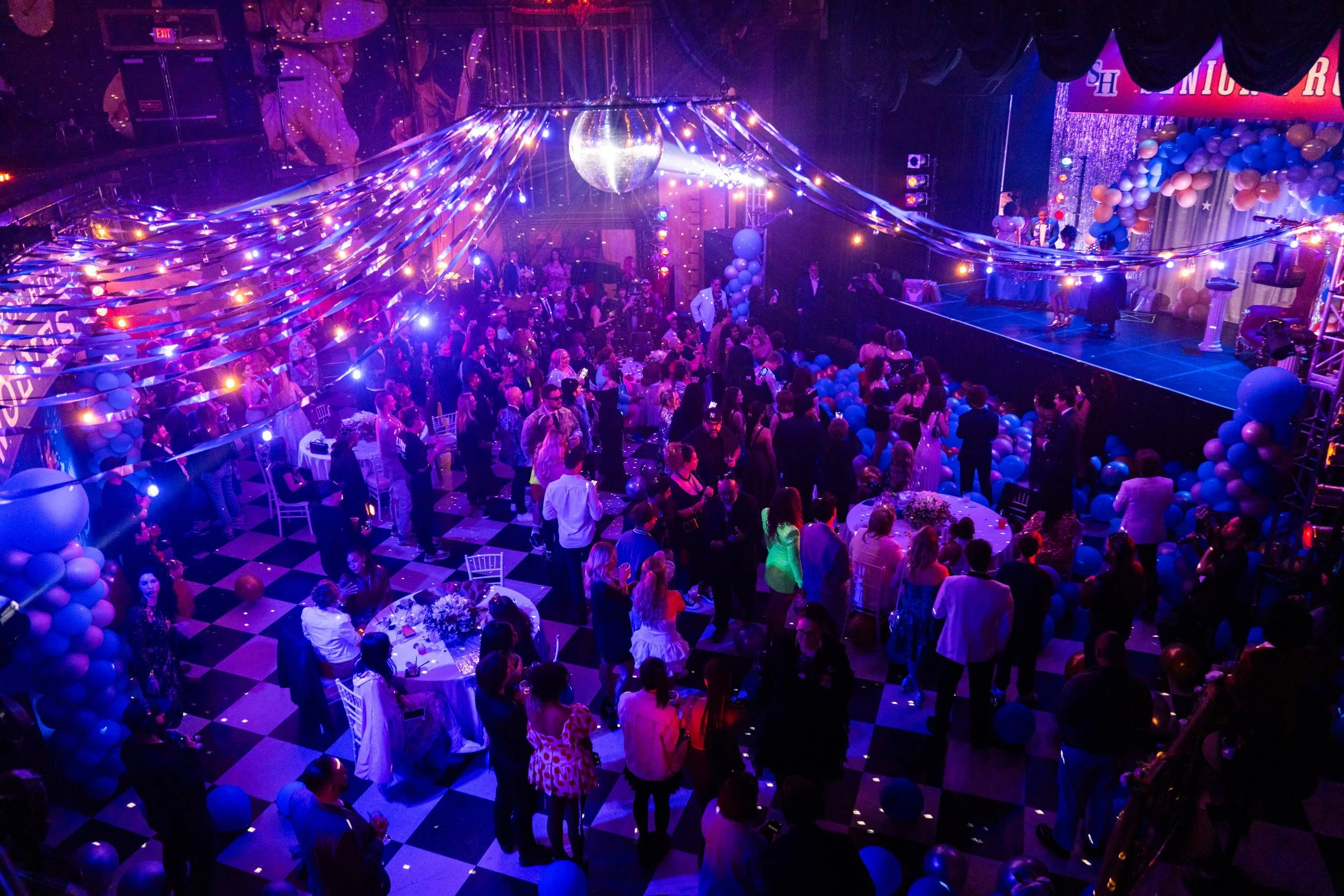 A large indoor party or celebration with many people dancing and socializing under colorful string lights, balloons, and a disco ball, with a stage on the right side.