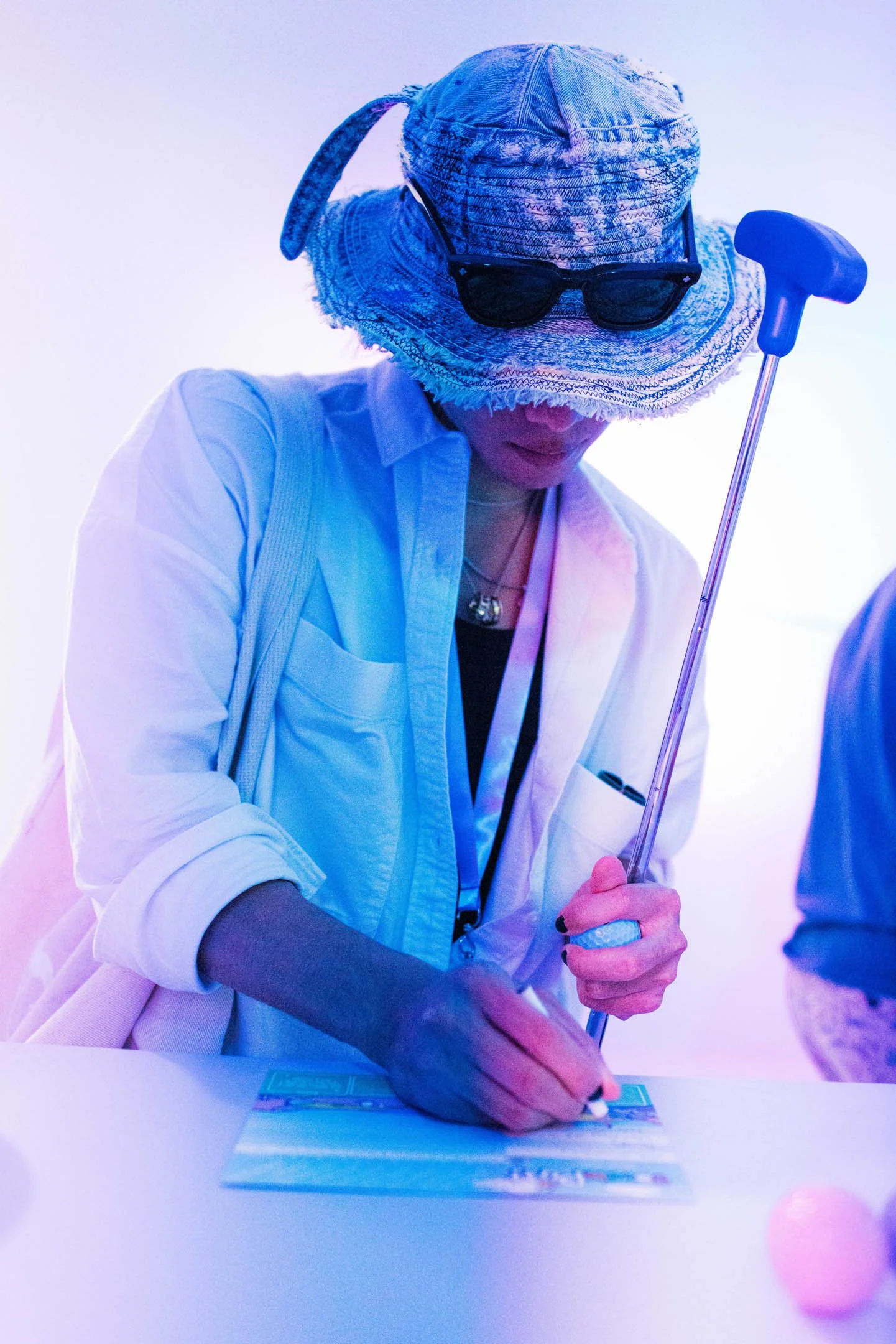 Person wearing a large wide-brimmed hat with sunglasses attached, writing on a card with a cane in hand, illuminated by blue and pink lighting.