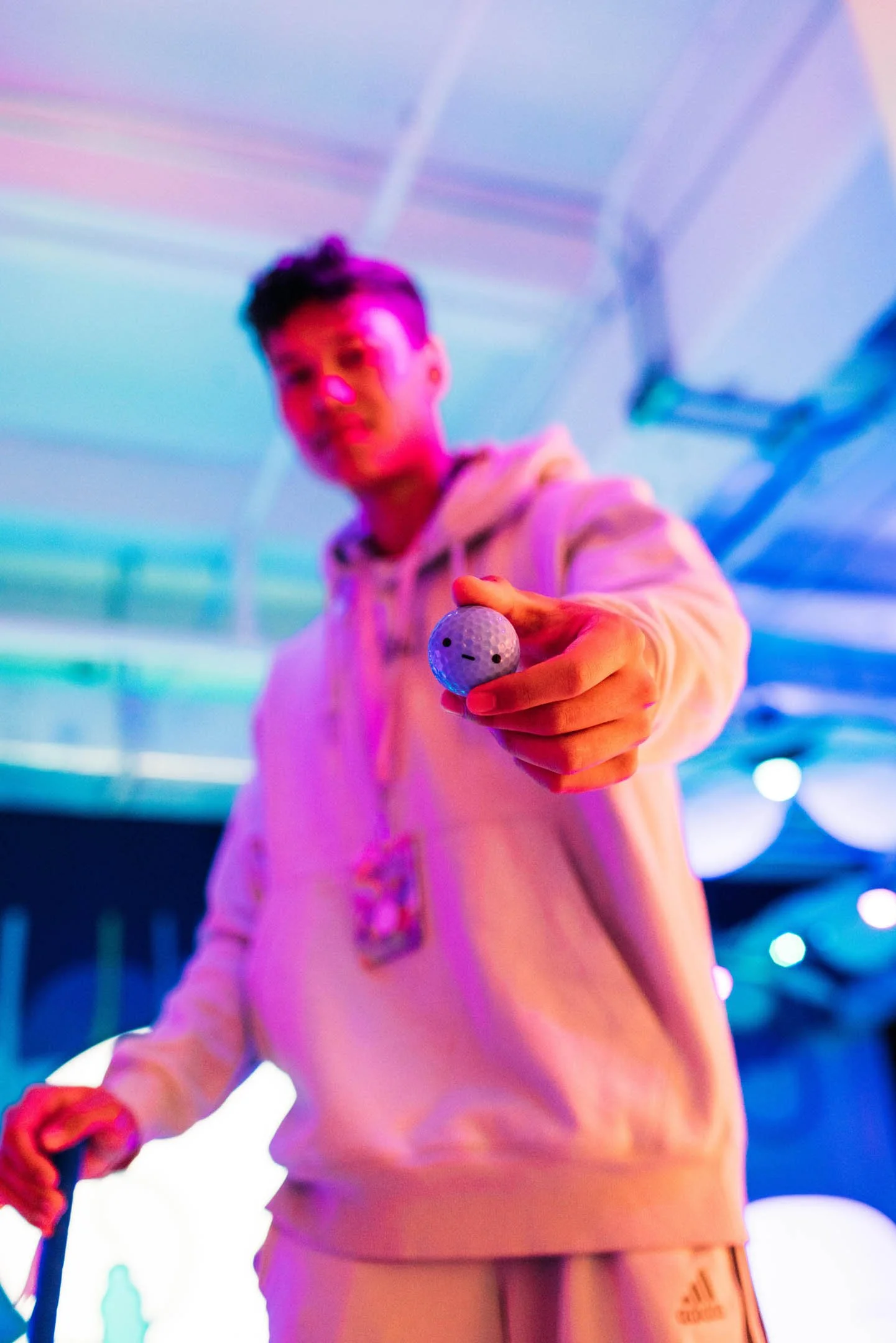 A young man holding a golf ball towards the camera with a blurred, colorful background.