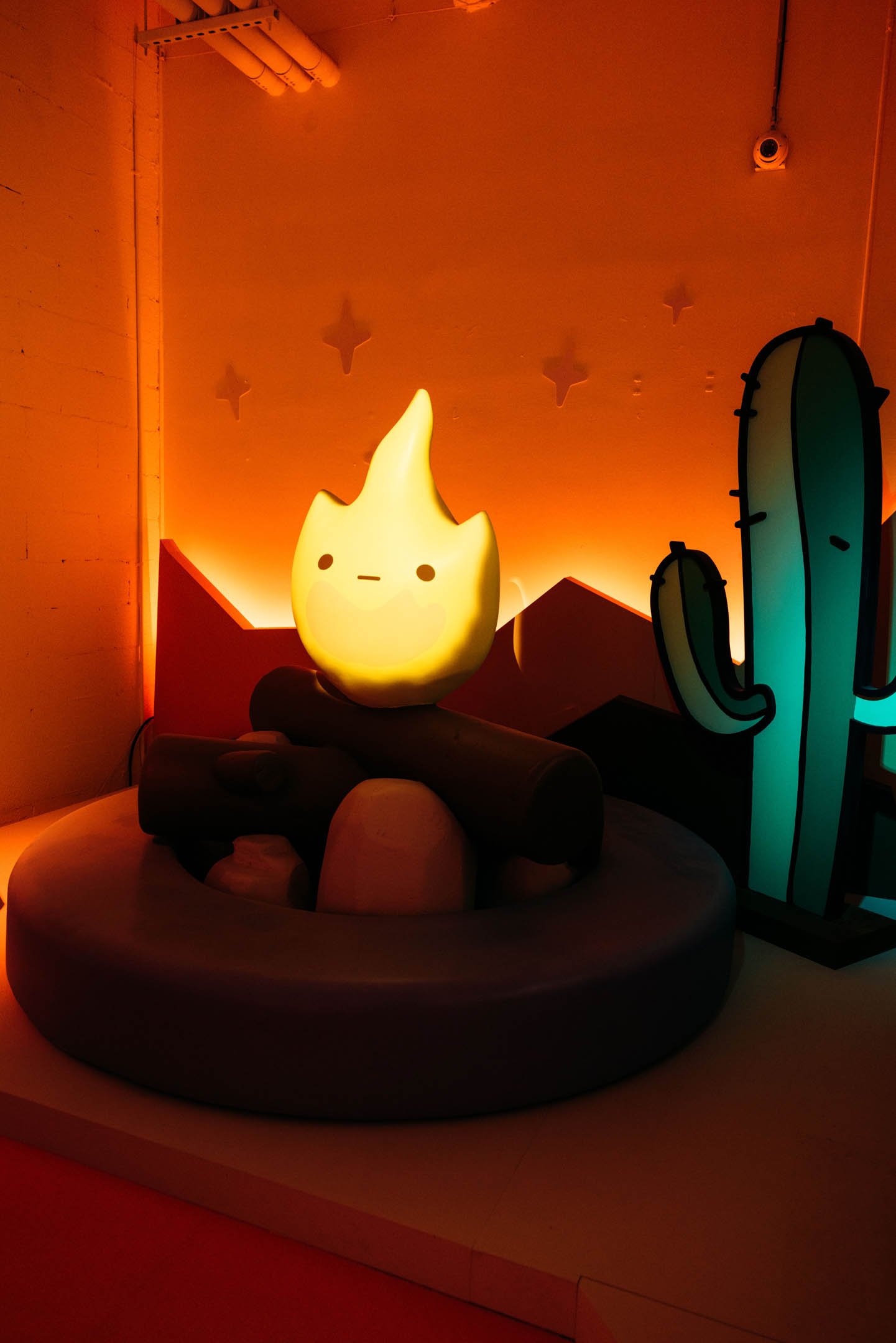 A glowing character resembling a cute, cartoon flame with a simple face, sitting on a log in an indoor setting with a desert-themed backdrop, including cacti and star decorations.
