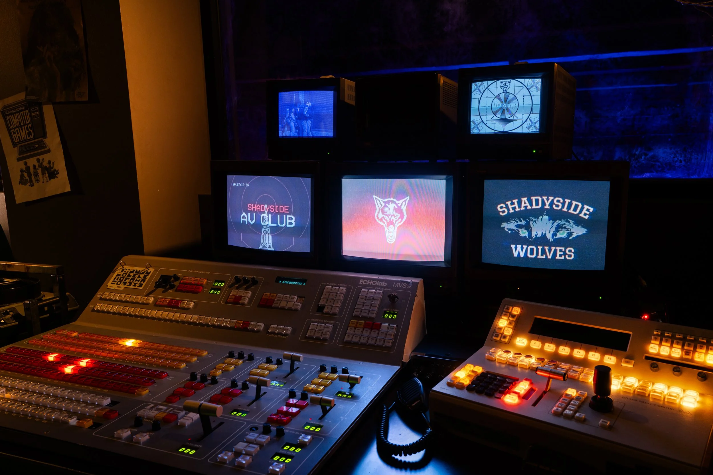 Multiple monitors displaying logos and graphics, along with a professional video control panel with various buttons and sliders in a dark studio setting.