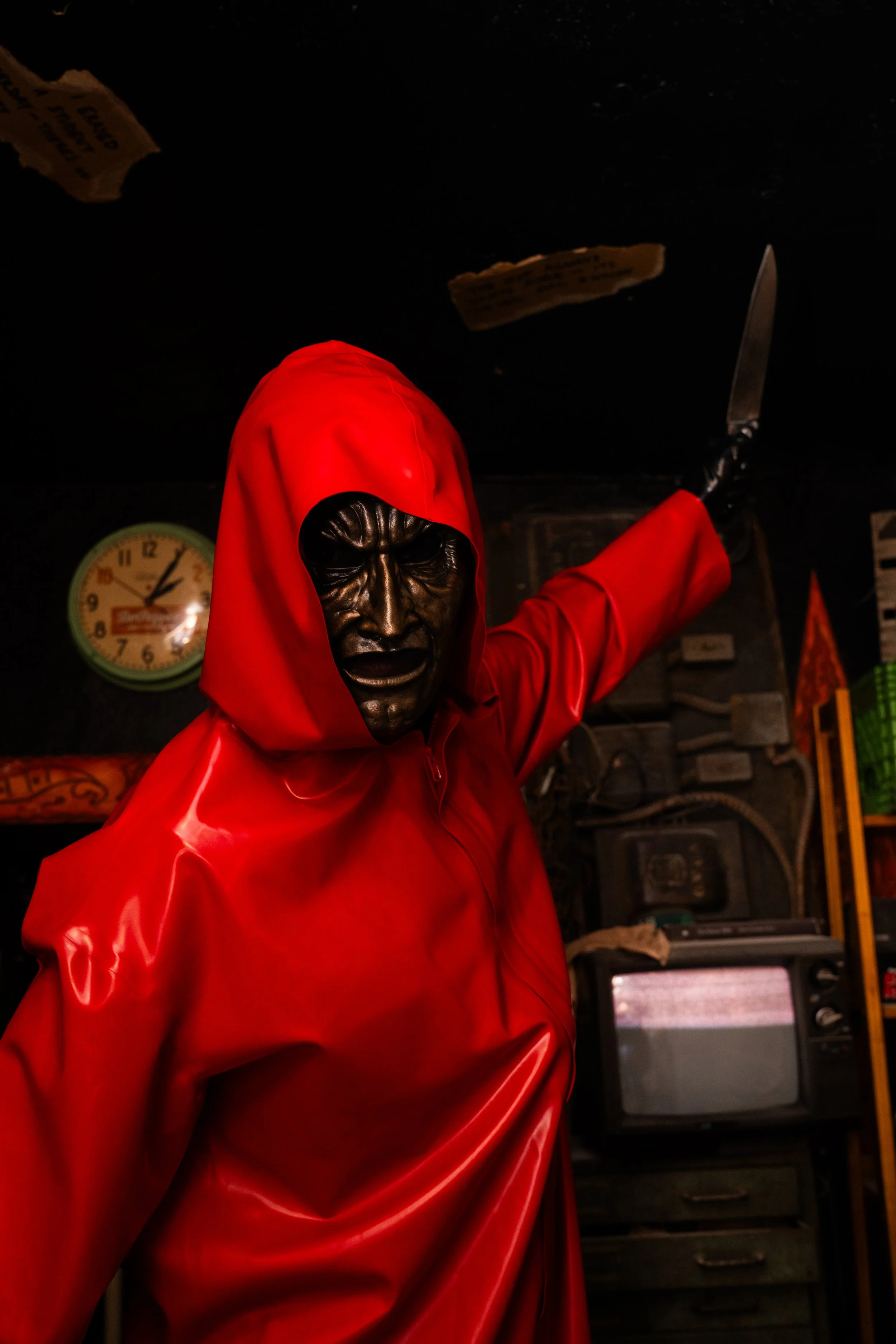 A person wearing a red hooded raincoat and a black mask with an angry face design, holding a knife, in a dark room with a clock on the wall and old electronic equipment.
