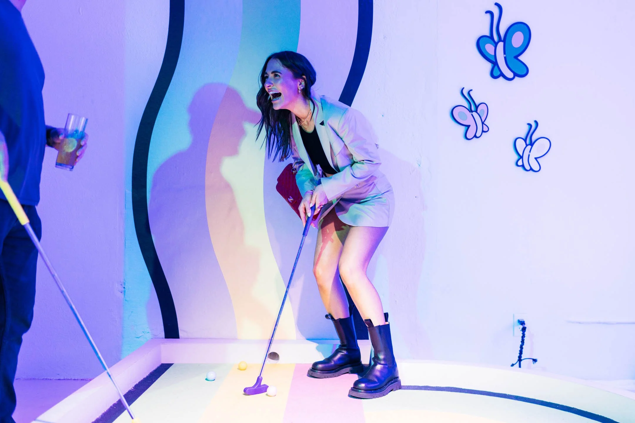 A woman playing mini golf in an indoor arcade with butterfly wall decorations, she is smiling and wearing a white blazer, black top, leather boots, and holding a golf club.