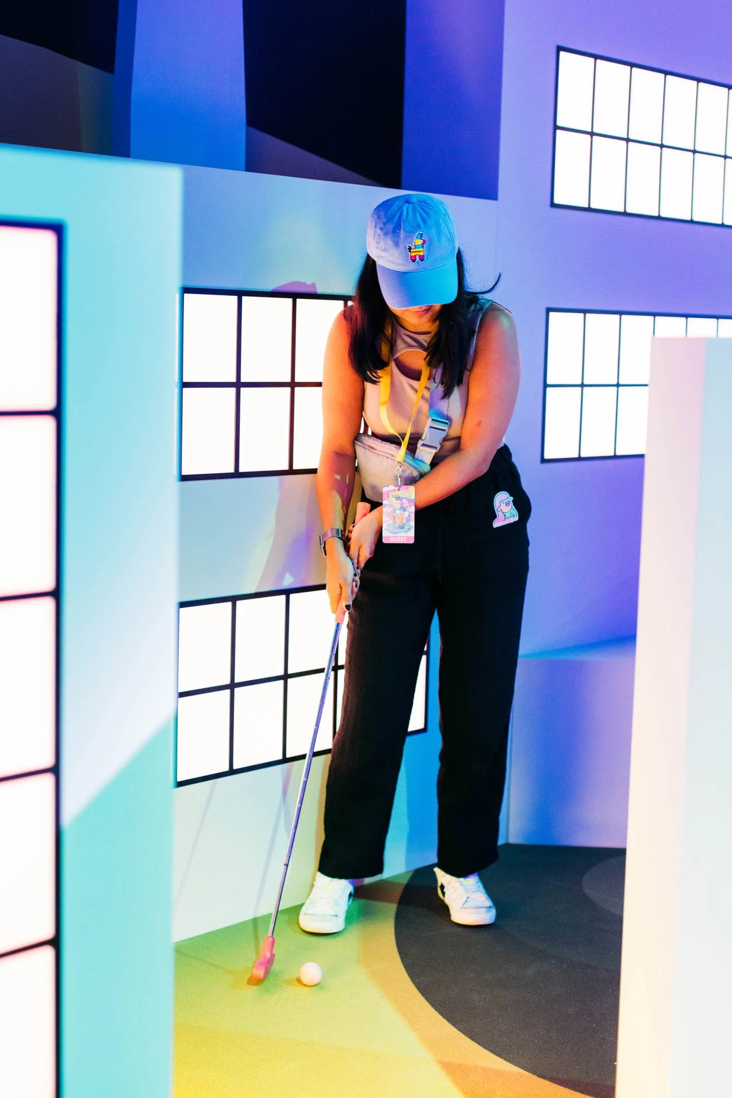 Woman dressed in casual attire playing mini golf, wearing a blue cap with an embroidered rainbow-colored gift box and a guest badge around her neck, guiding her golf shot on a colorful indoor course with illuminated window-like panels.