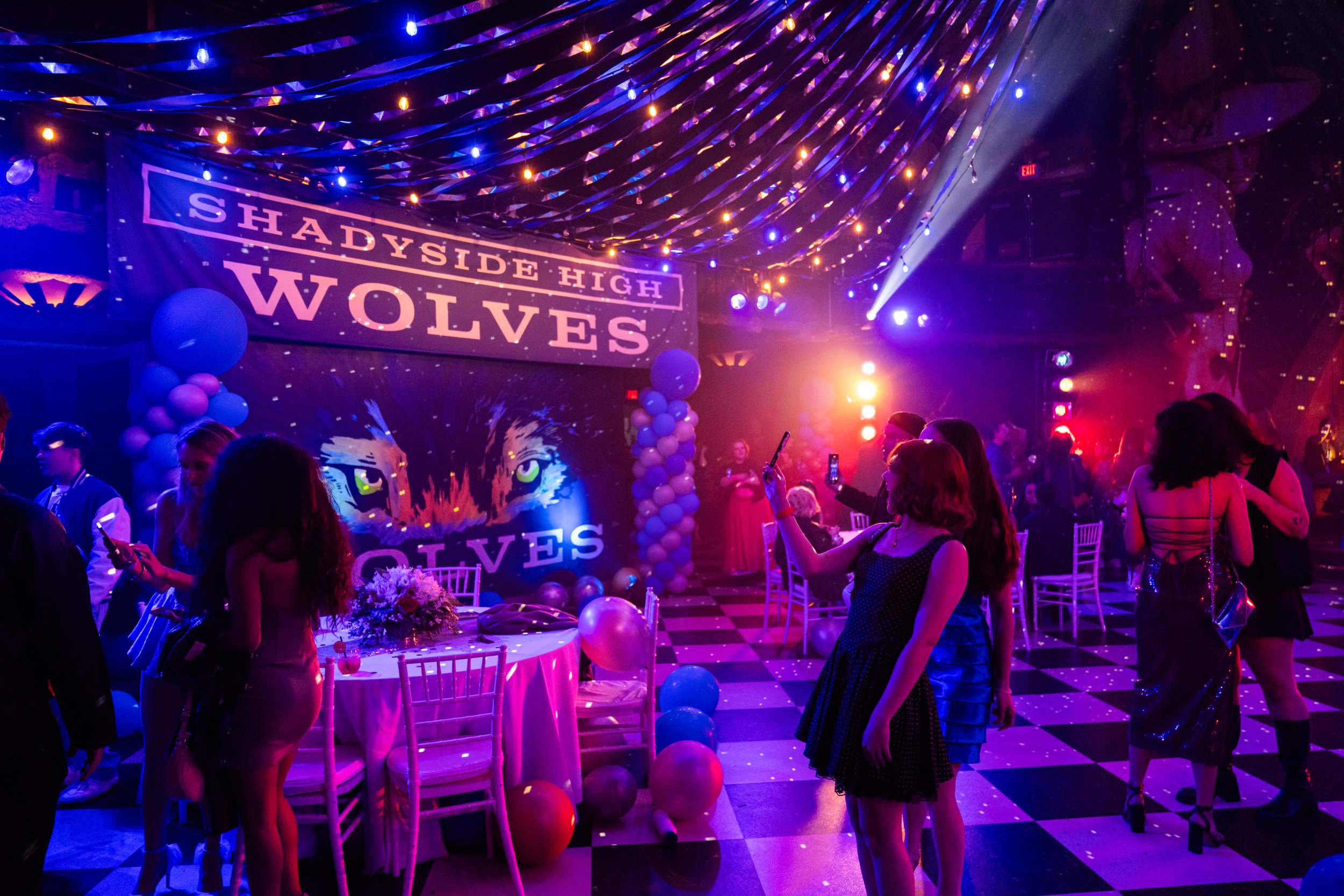 People dancing and taking photos at a party with a banner reading 'Shadyside High Wolves,' balloons, and colorful lighting.