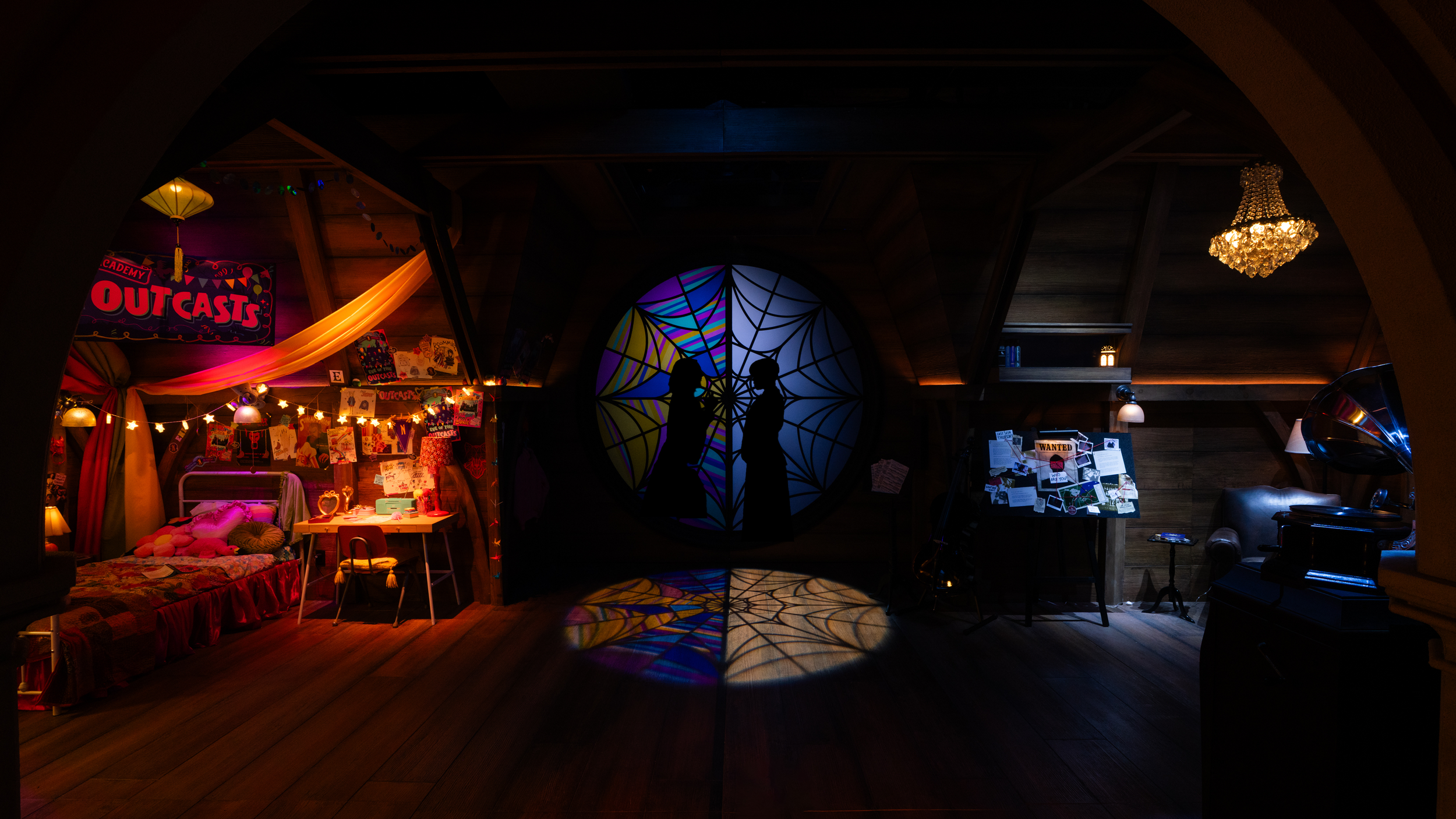 Dark room with a silhouette of a couple in front of a stained glass window depicting spiderwebs, illuminated with colorful lights. To the left, a decorated bedroom scene with a bed, stuffed animals, and fairy lights. To the right, a bulletin board wi