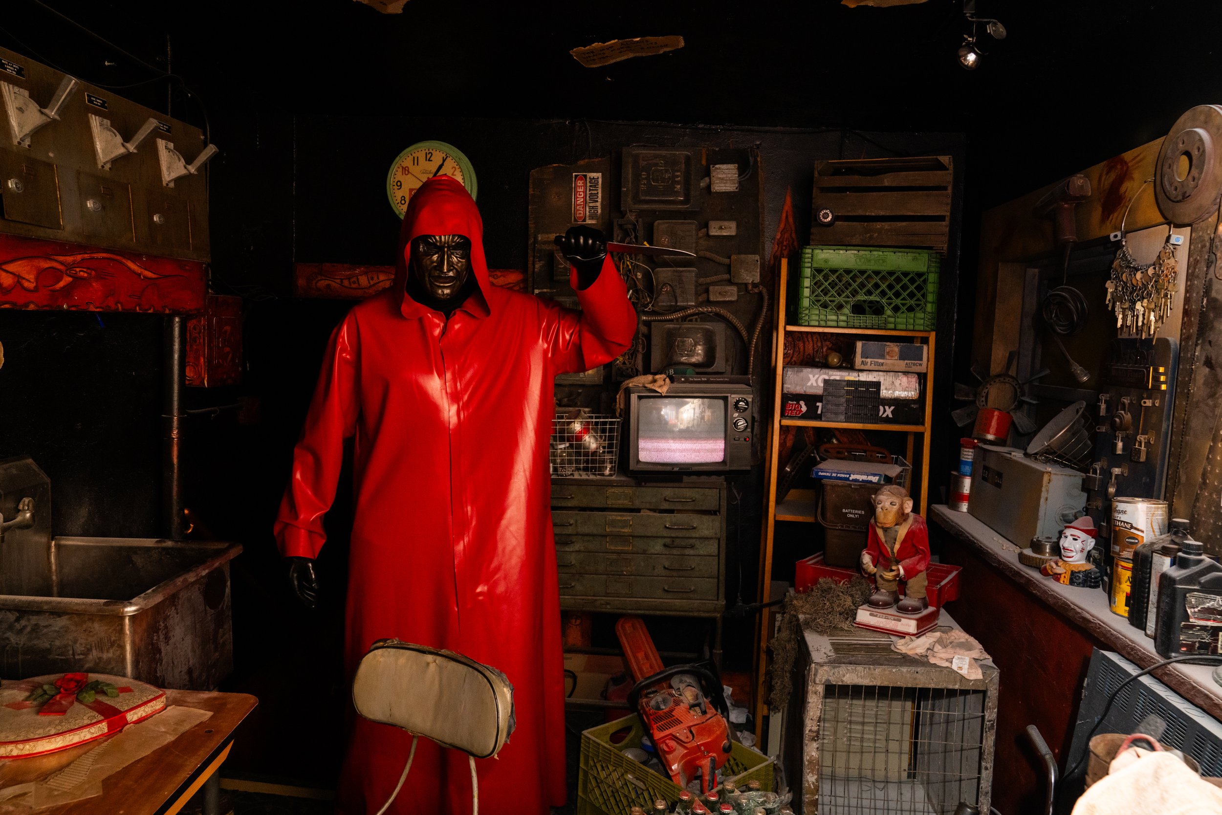 A person wearing a red hooded raincoat and black gloves stands in a cluttered room, holding a knife. The room is filled with old electronics, toys, and miscellaneous items, with dark walls and a clock on the wall.
