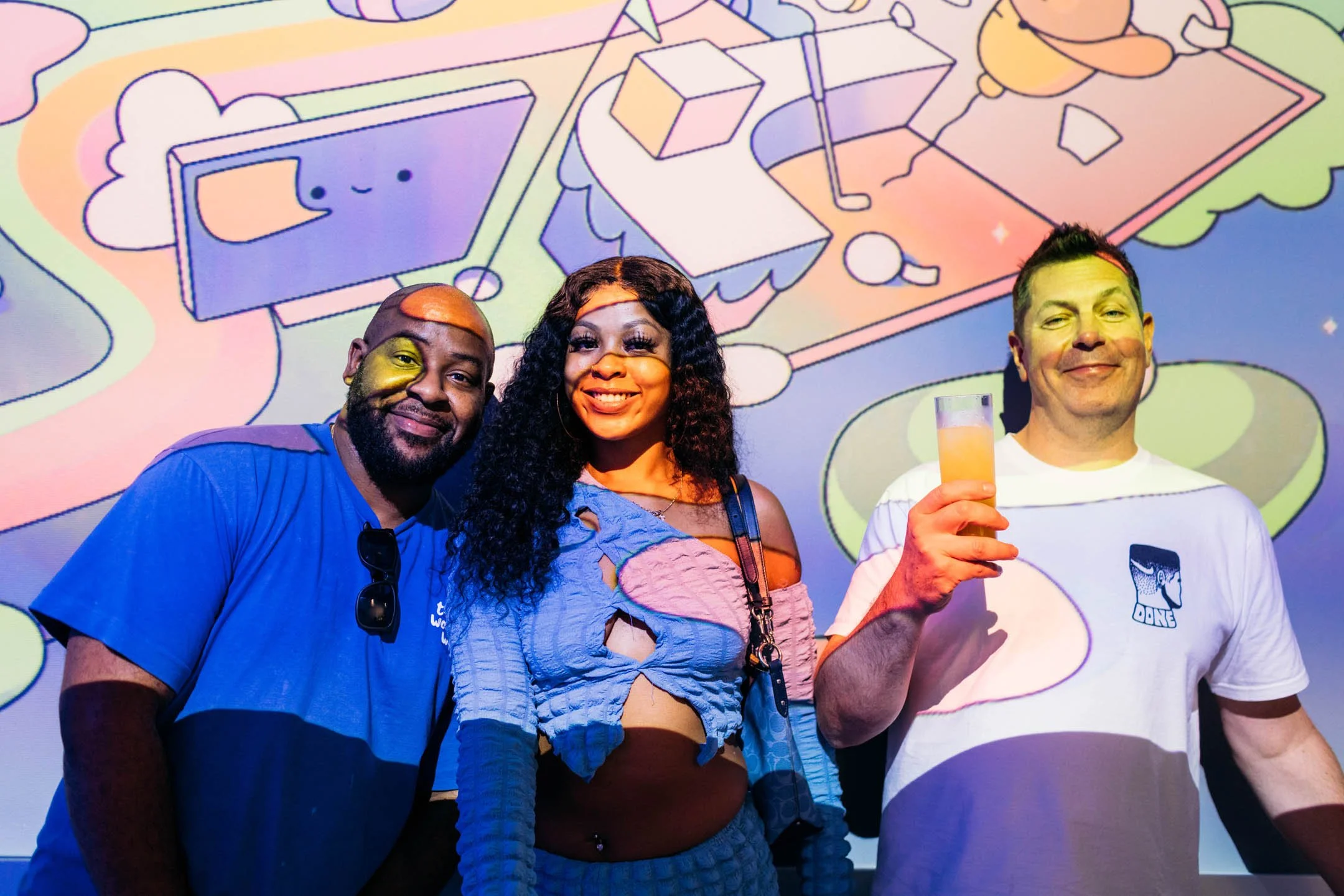 Three people smiling at a party with a colorful animated background projected behind them. The person on the left is a man wearing a blue shirt and sunglasses hanging from his collar. The person in the middle is a woman with long curly black hair, we