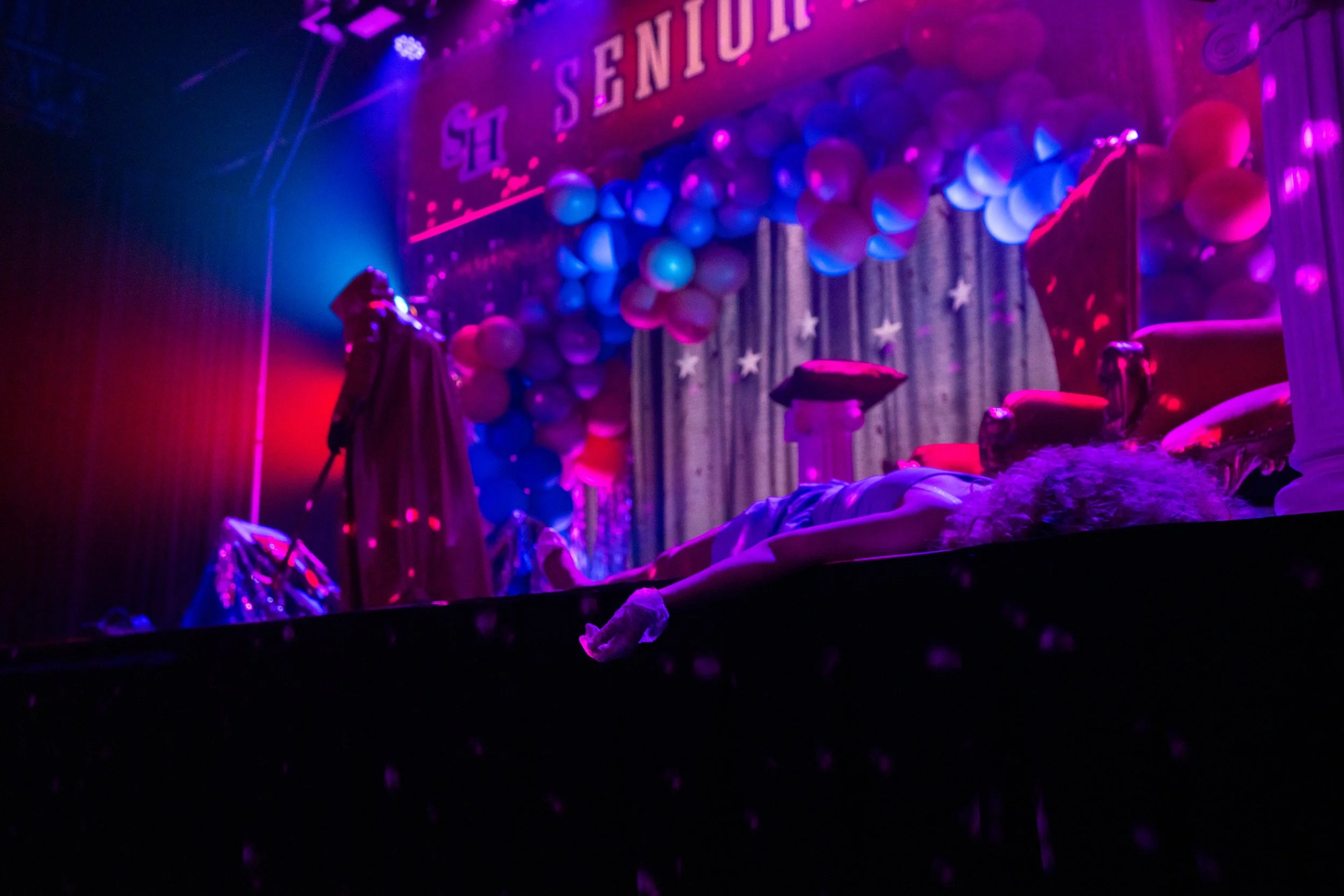 A theatrical stage decorated with balloons, velvet curtains, and ornate chairs, lit with purple and blue stage lights. A person is lying on the stage with curly blonde hair, while another person in a cape and hat stands nearby. The background feature