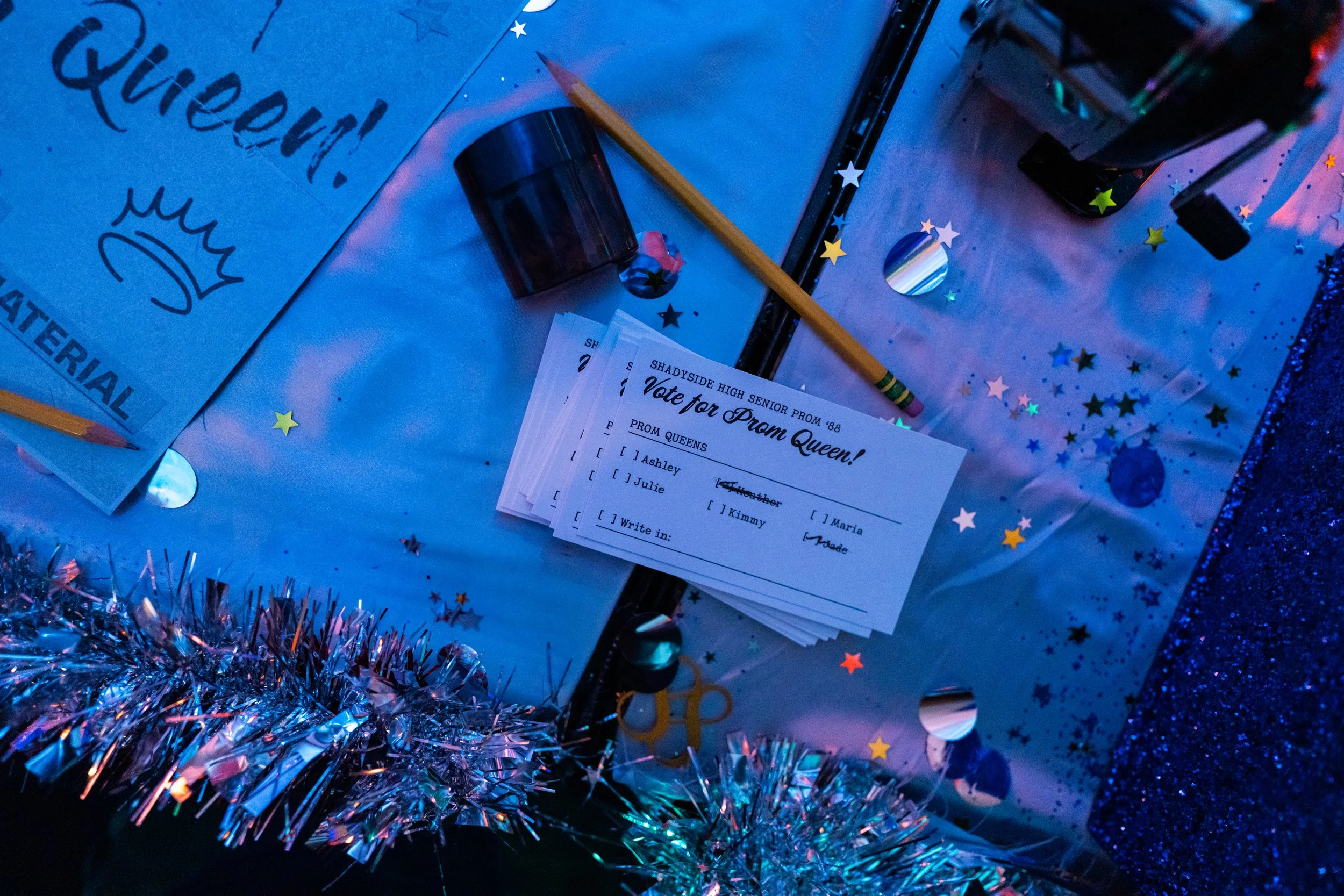 A prom king or queen vote sheet with options Ashley, Julie, Kimmy, Maria, and Wade, placed on a decorated table with star-shaped confetti, tinsel, pencils, a marker, and a roll of tape.
