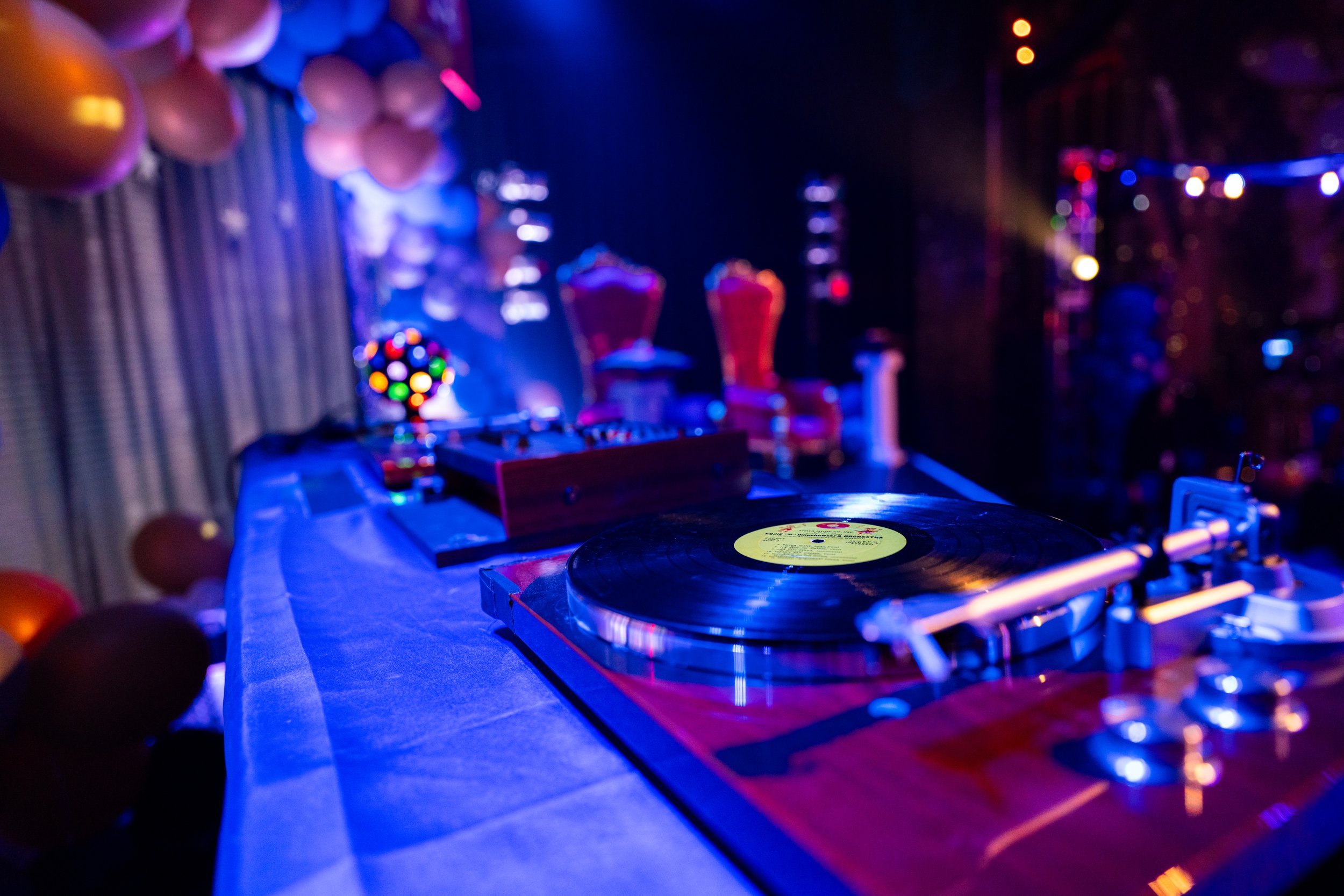 A DJ turntable and mixer on a table, with colorful party decorations and balloons in the background, set in a dimly lit party or nightclub scene.