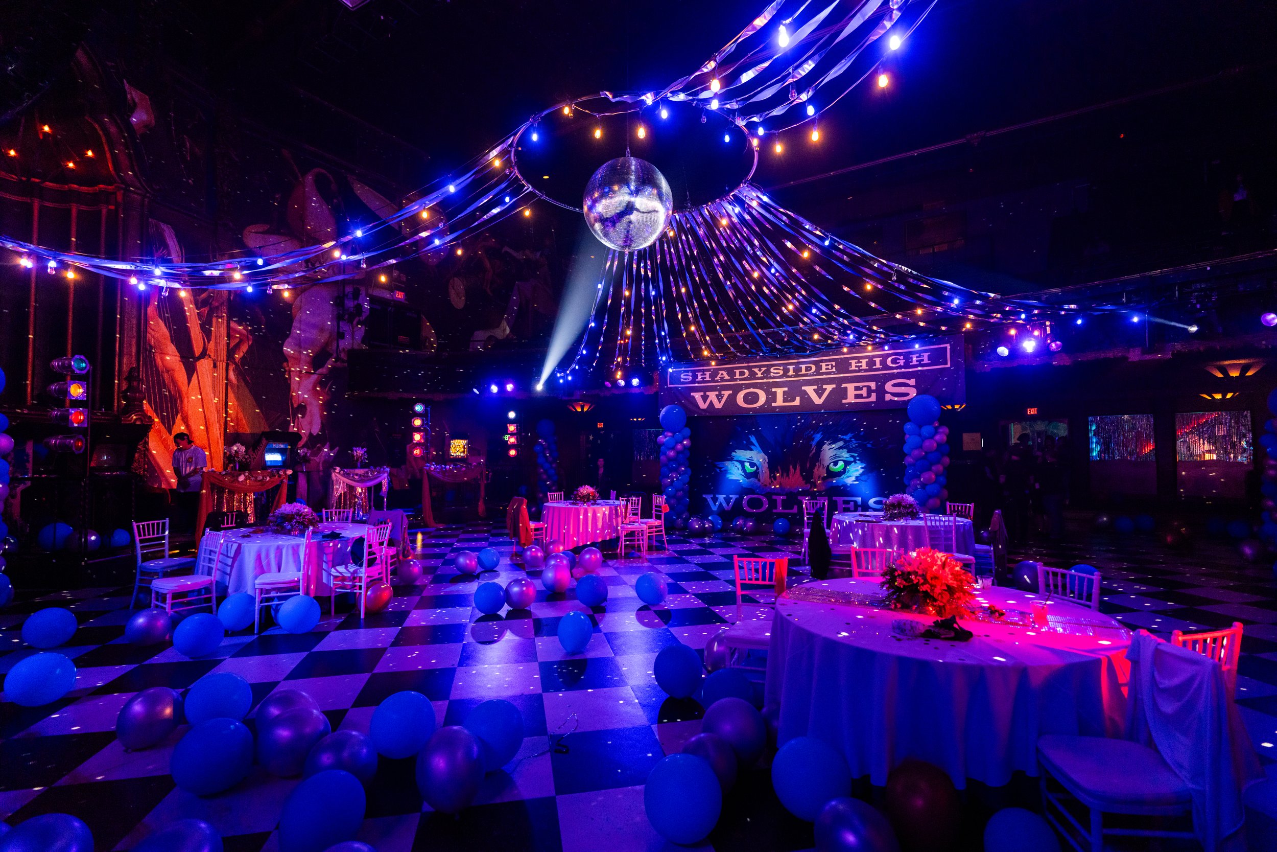 Party decorated with blue and purple lighting, balloons, and a dance floor. Large banner reads 'Shadyside High Wolves' with an image of a wolf's face.