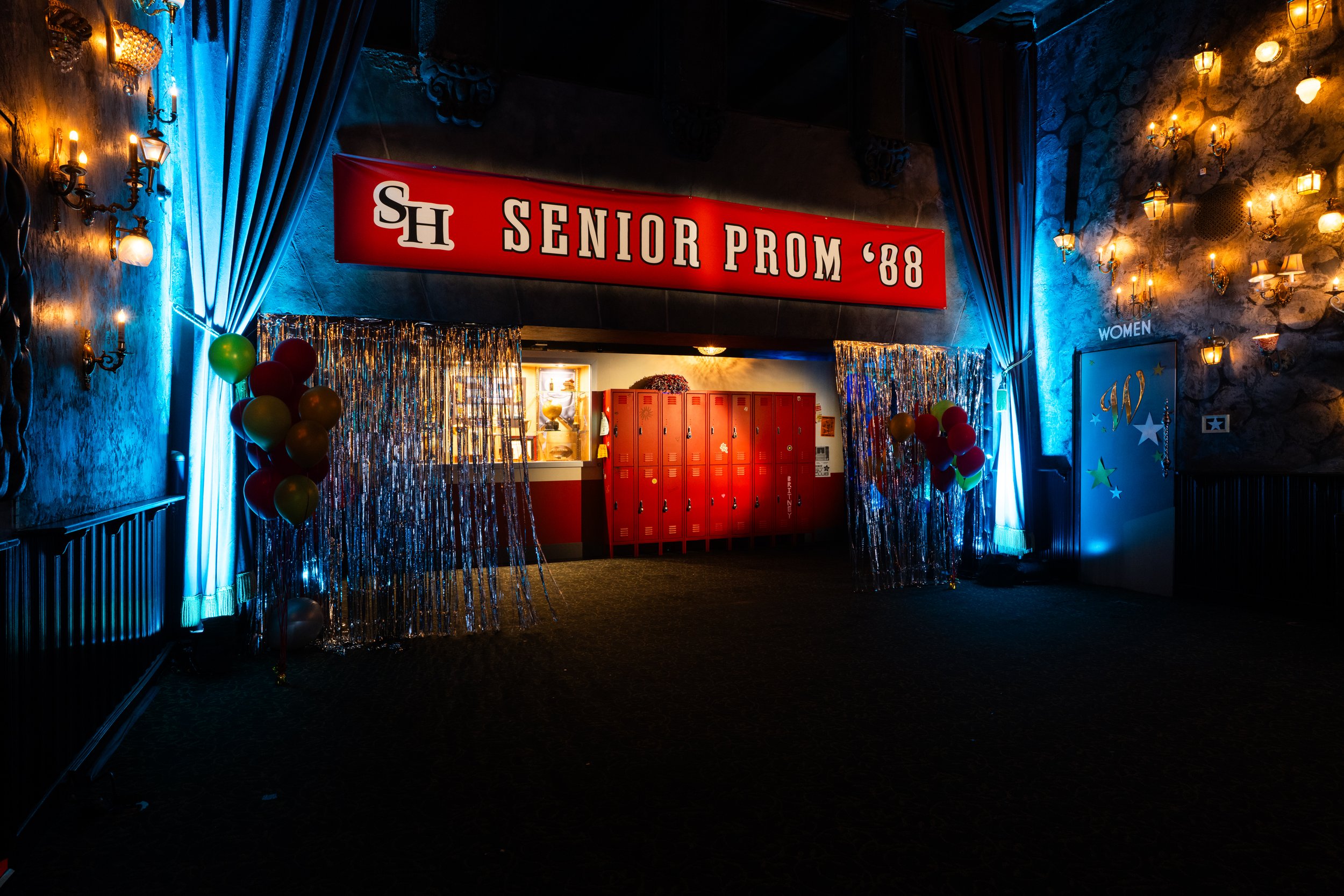 Decorated entrance to a senior prom event with a red banner reading 'SH Senior Prom ’88', lockers, balloons, curtain dividers, and ambient lighting