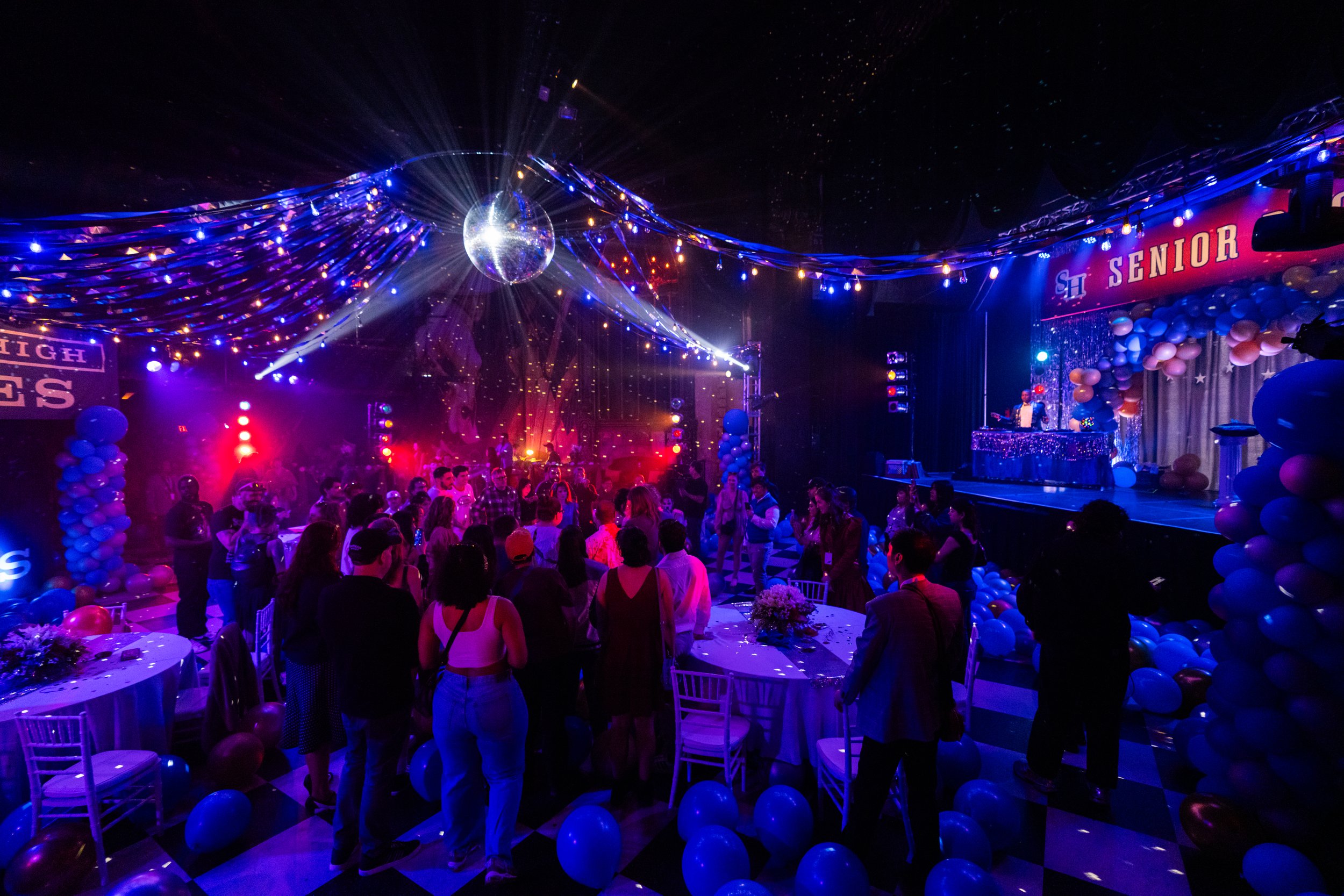 A crowded school prom dance with blue and purple lighting, balloons, a disco ball at the top center, and a decorated stage with a sign that says 'Senior'.