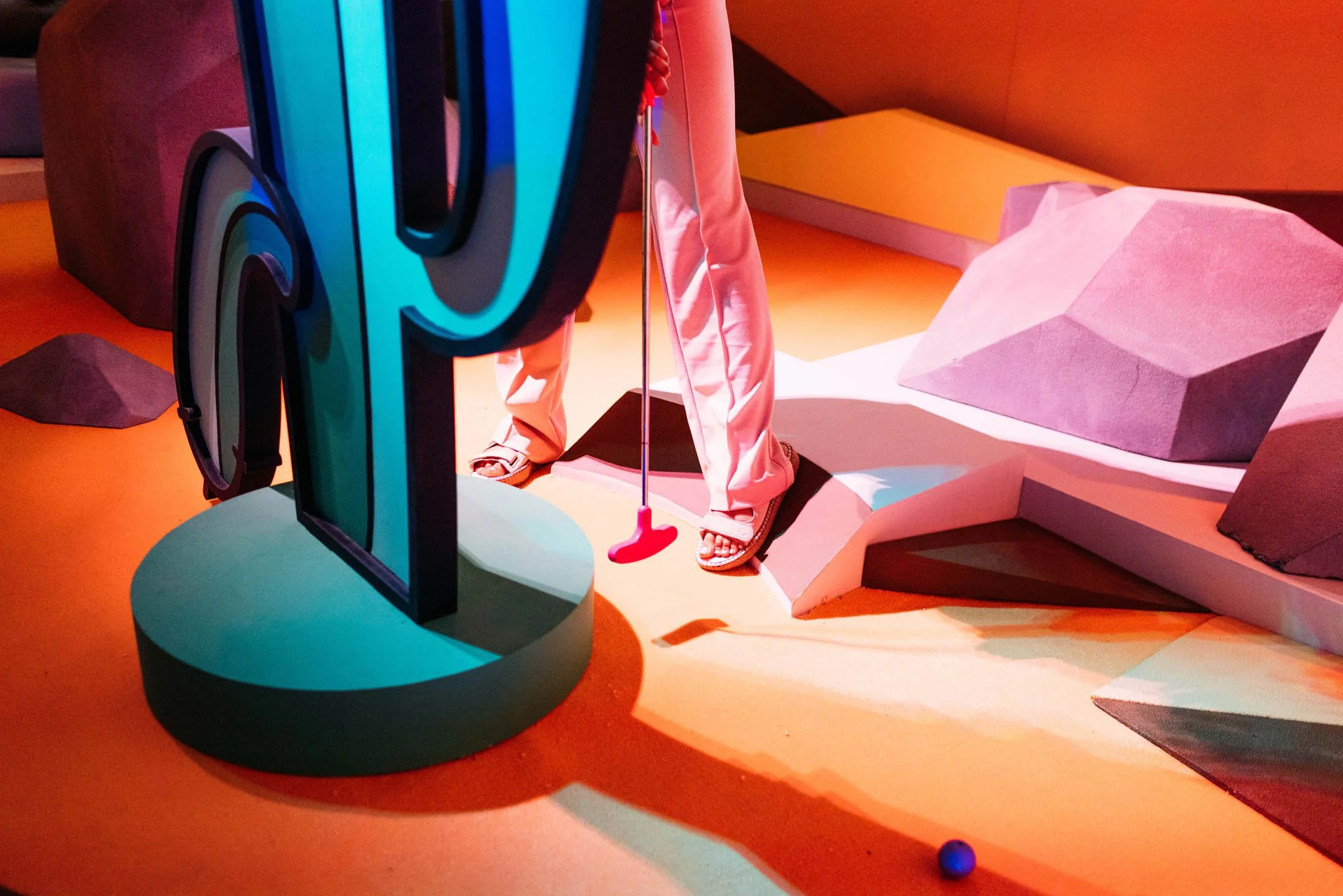 Part of a colorful abstract art installation with a person in white pants and sandals holding a golf club, surrounded by geometric shapes and bright lighting.