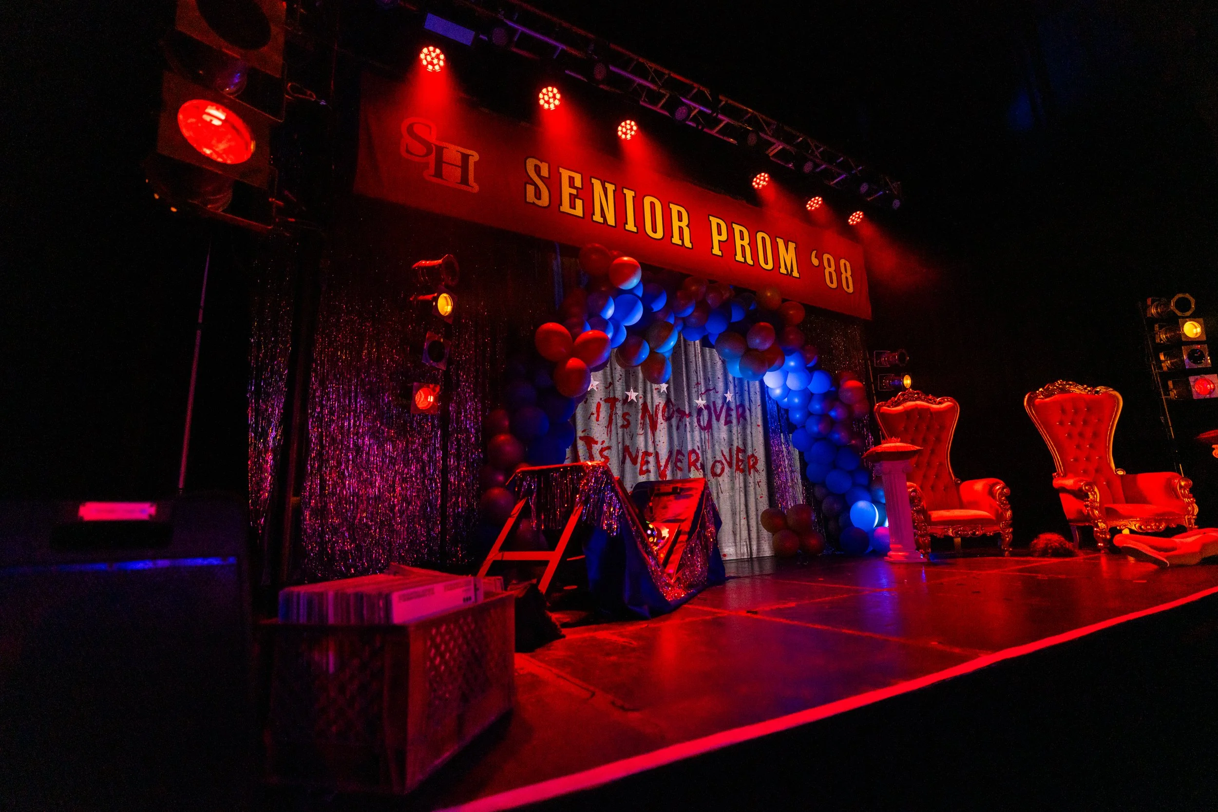 Stage decorated for a senior prom with a red banner reading 'Senior Prom '88', inflatables, balloons, glitter curtains, and two ornate red chairs.