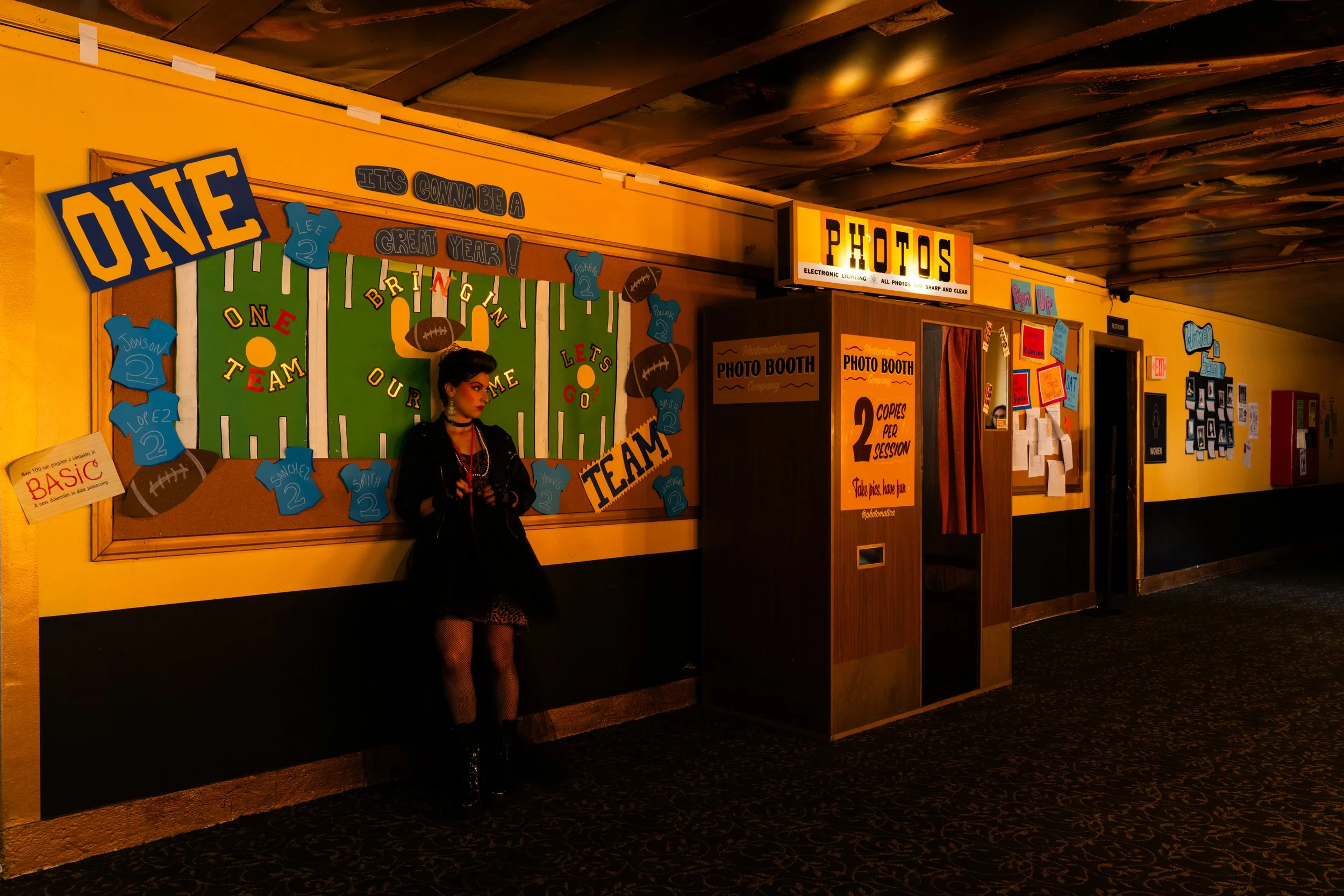A girl standing in a sports-themed hallway near a photo booth, decorated with football-themed posters, signs, and wall art, with a scoreboard backdrop and a yellow wall.