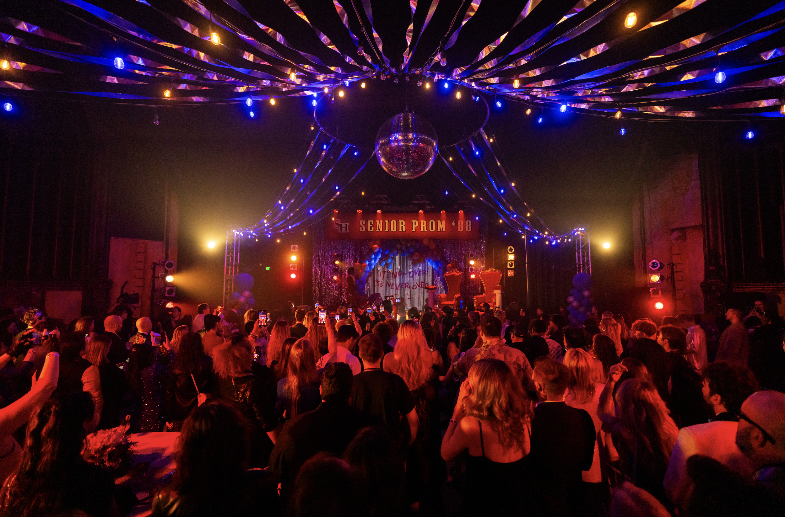 A large indoor celebration event with many people attending a senior prom in 1988. The stage has a sign that reads 'Senior Prom '88,' decorated with balloons, silver fringe curtains, and giant chairs, with vibrant colorful lighting and a disco ball h