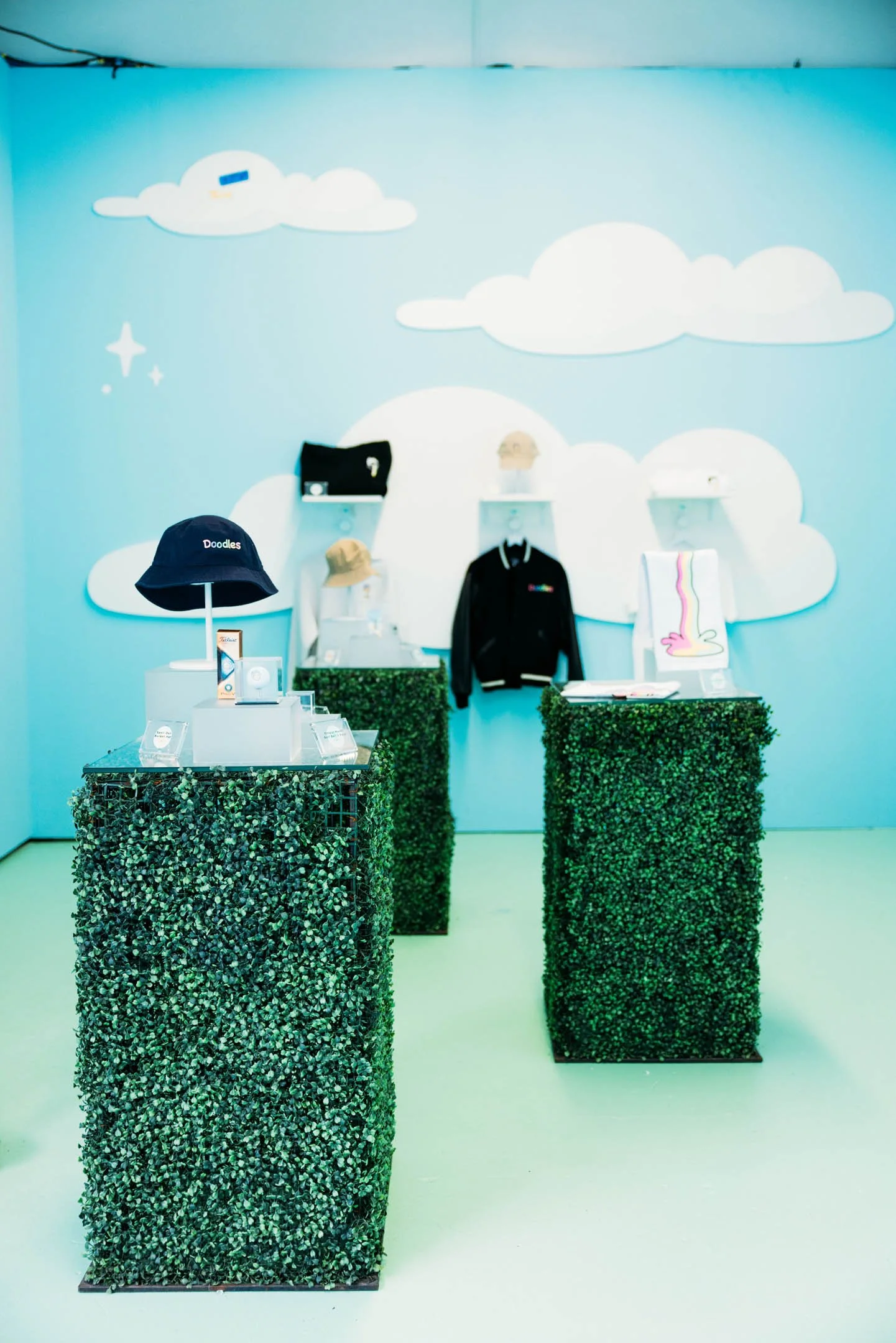 A retail display area with two green hedge-covered pedestals showcasing various items, including hats, a black jacket on a display hanger, and other accessories, set against a light blue wall decorated with clouds, a rainbow, and stars.