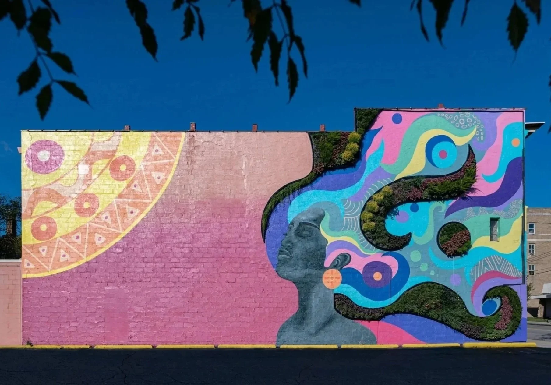 Colorful mural on a brick wall depicting a profile of a woman's face with flowing, abstract hair in vibrant colors, with a yellow sun in the upper left corner and a pink background.