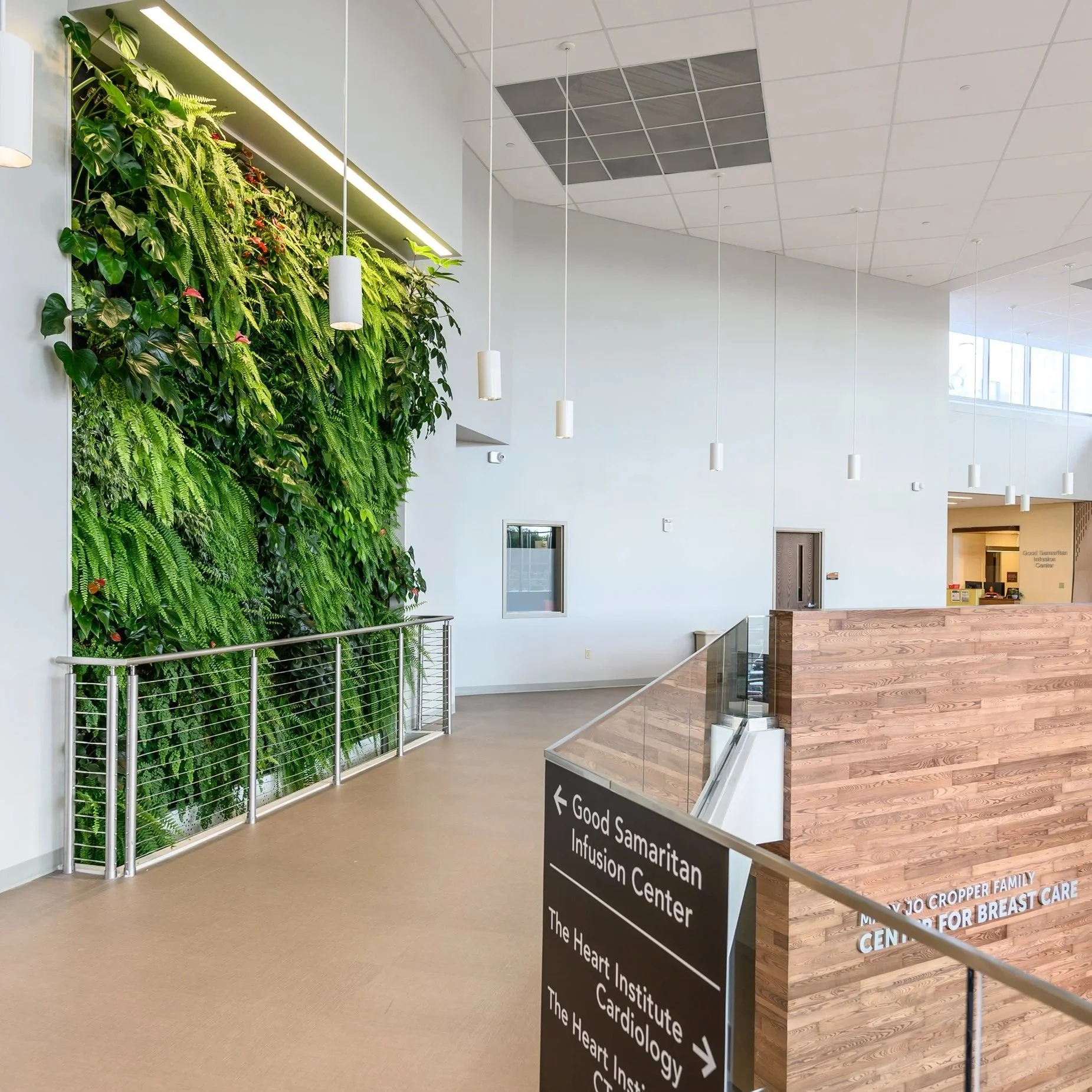 Interior of a medical facility lobby with a green wall of plants, hanging lights, and directional signage for various health centers.