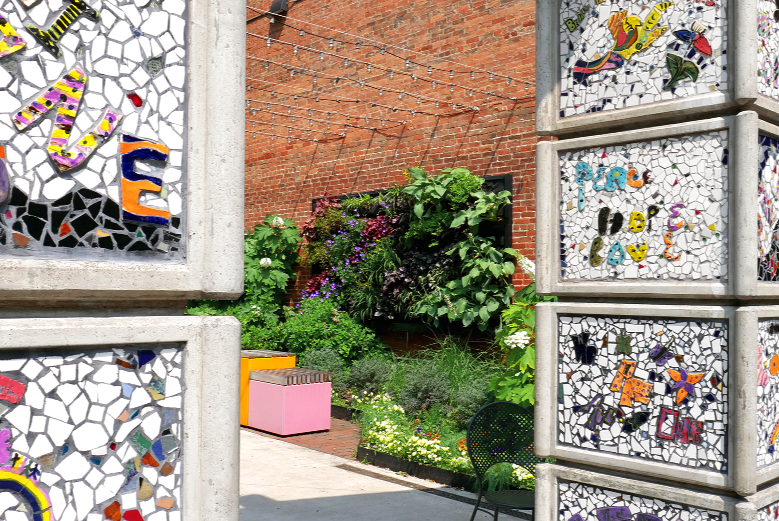 Colorful mosaic art panels along with designs of animals and flowers, partially framing a garden with green plants and a lush, vibrant living wall with colorful flowers, a pink box, and outdoor chairs against a brick wall and string lights.