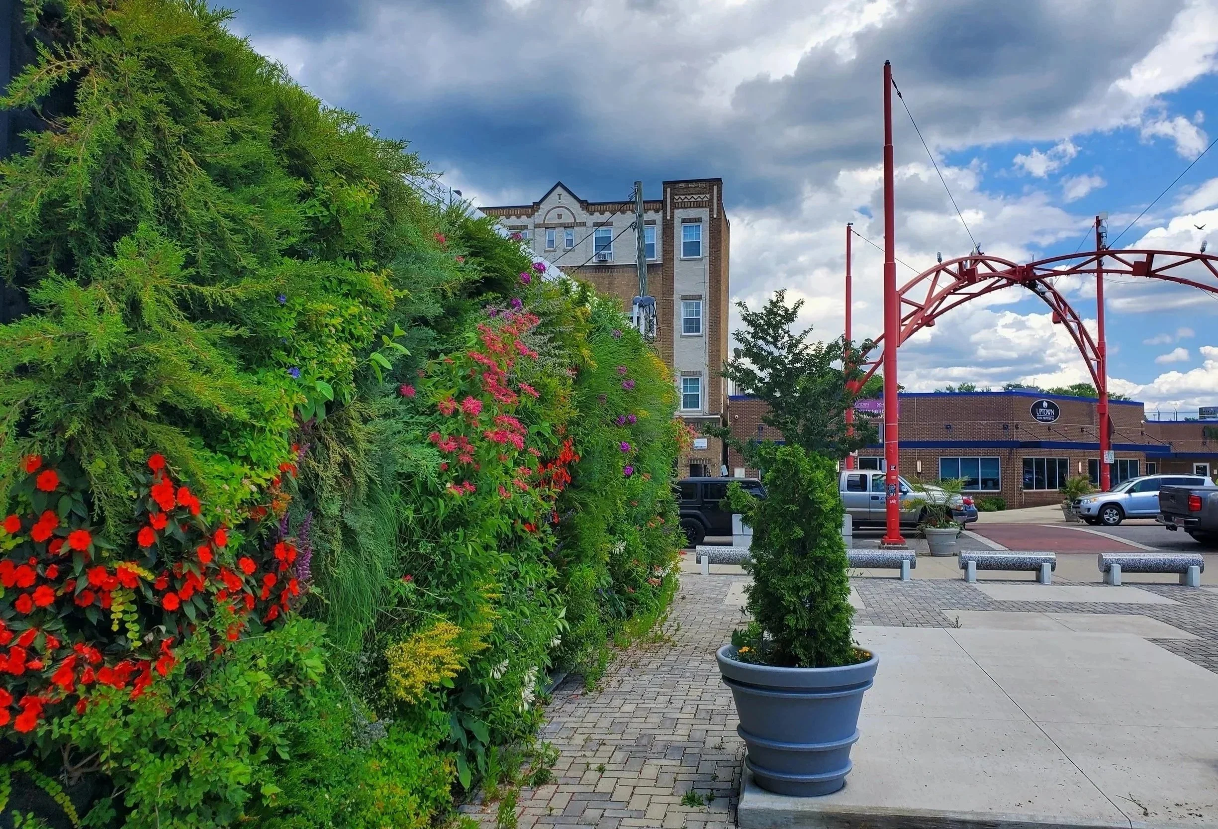 A sidewalk with a colorful flower wall on the left, a small potted tree in the foreground, parked cars, a building with signs, and a red arched structure overhead under a partly cloudy sky.