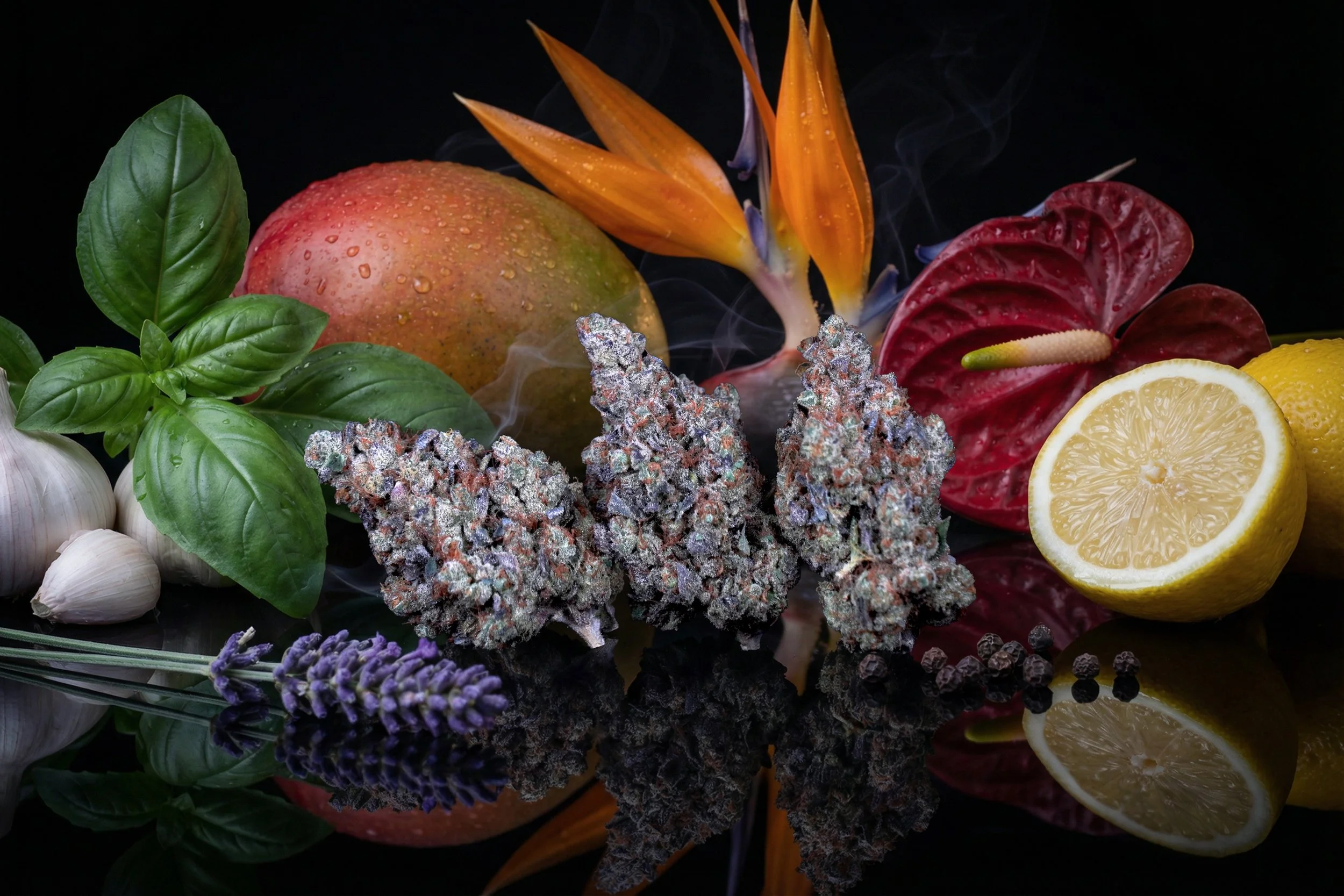 A collection of various fresh ingredients, including an apple, basil leaves, garlic cloves, lavender, cannabis buds, a lemon, a red anthurium flower, a bird of paradise flower, and black peppercorns, arranged on a reflective black surface.