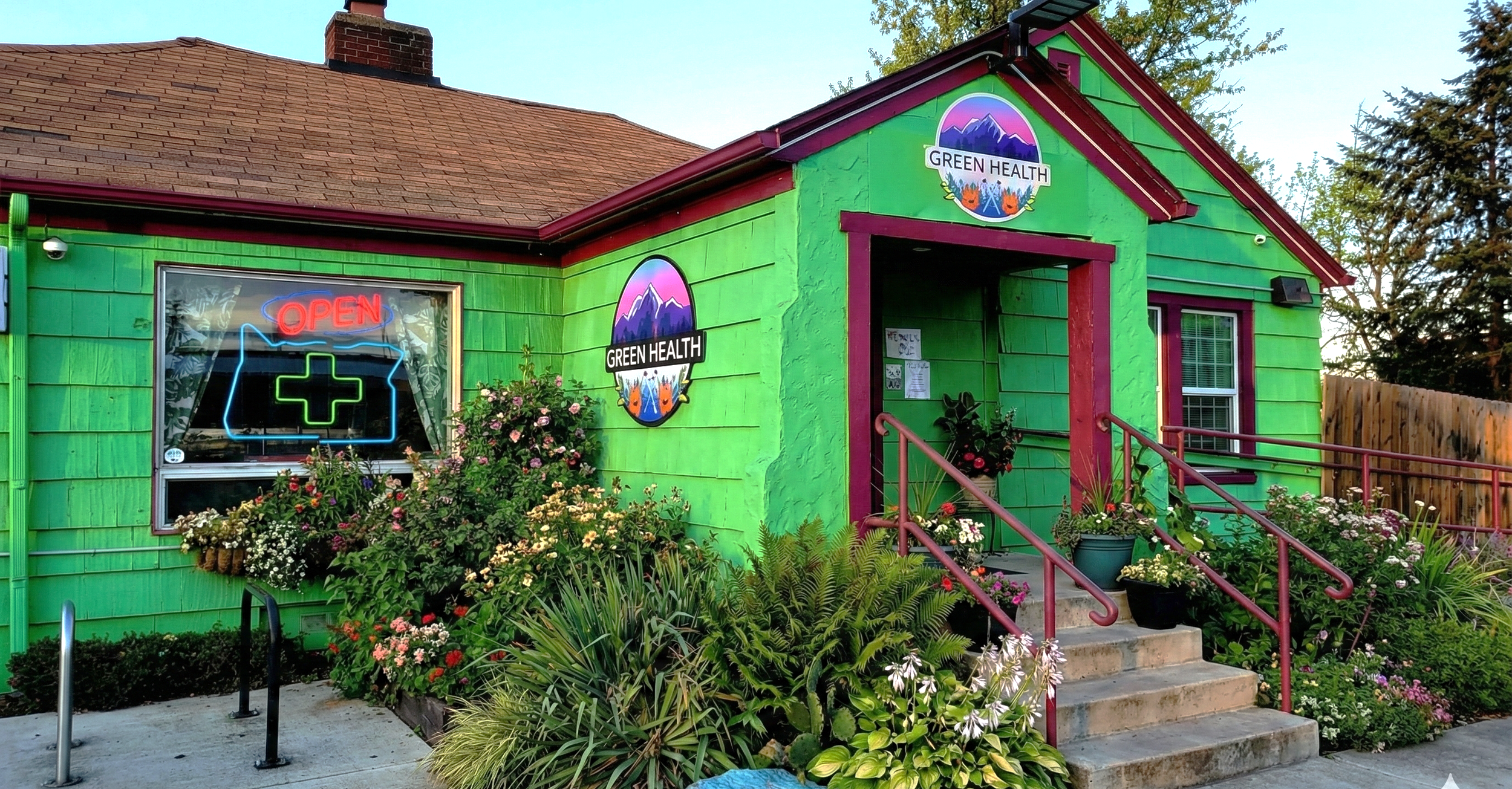 A colorful building painted bright green with purple trim, with signs indicating it is a healthcare clinic called 'Green Health.' The building has flower beds with various flowers in front, a staircase with pink handrails leading to the entrance, and a neon sign in the window that says 'Open' with a medical cross. Mountain logos are on the signs, and there is a wooden fence and trees in the background.