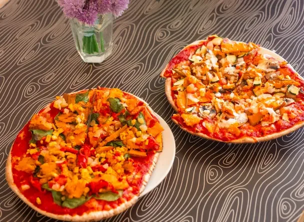 Two vegetable pizzas on a Night Kitchen table beside a vase of flowers.
