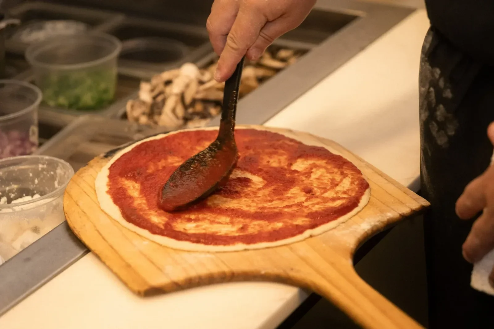 Tomato sauce being spread over fresh pizza dough on a wooden peel at Night Kitchen.