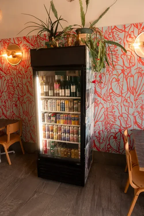 Glass-front beverage fridge stocked with bottles and cans beside a colourful wall at Night Kitchen.