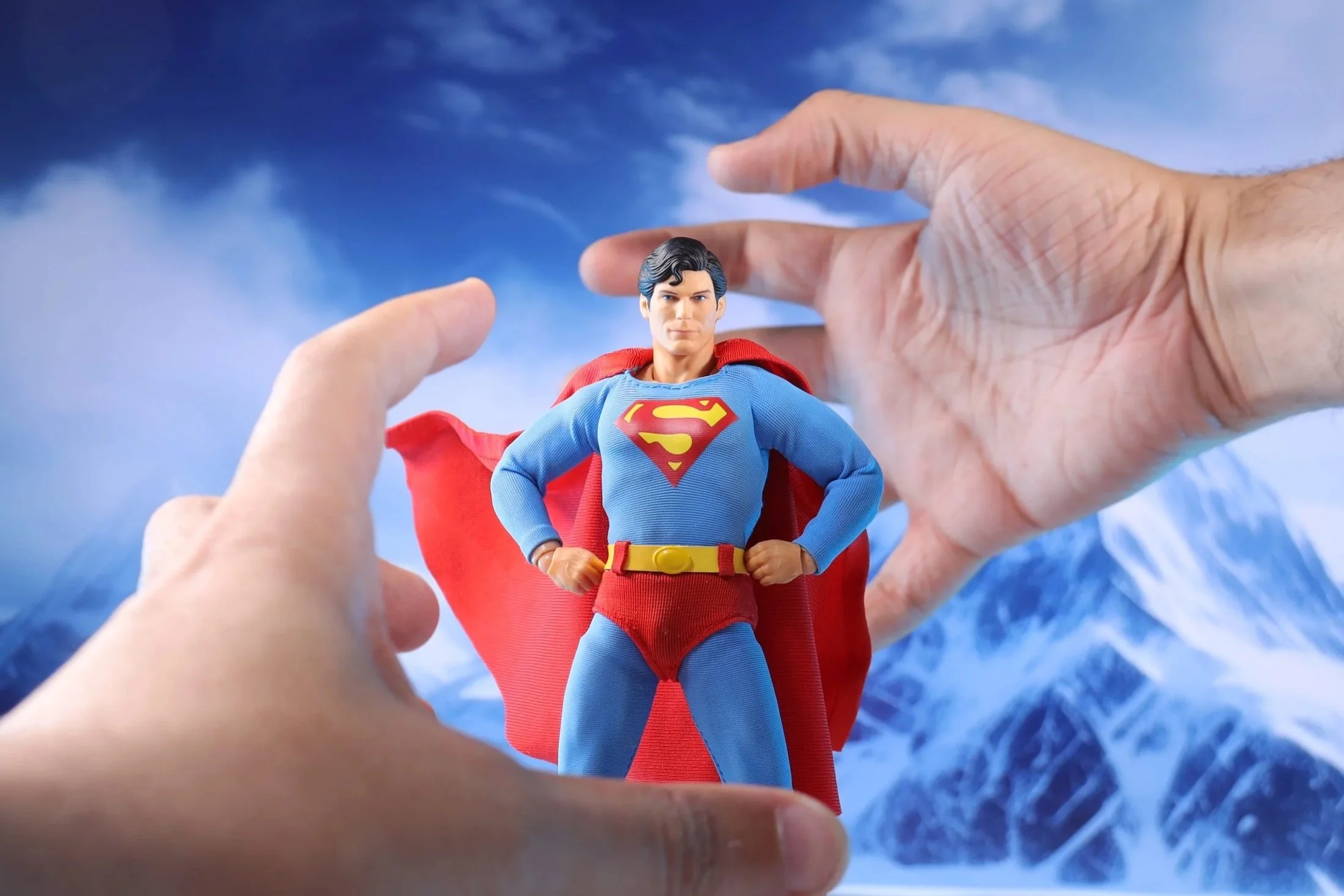 A person's hands holding a Superman action figure against a background of a snowy mountain landscape and blue sky.