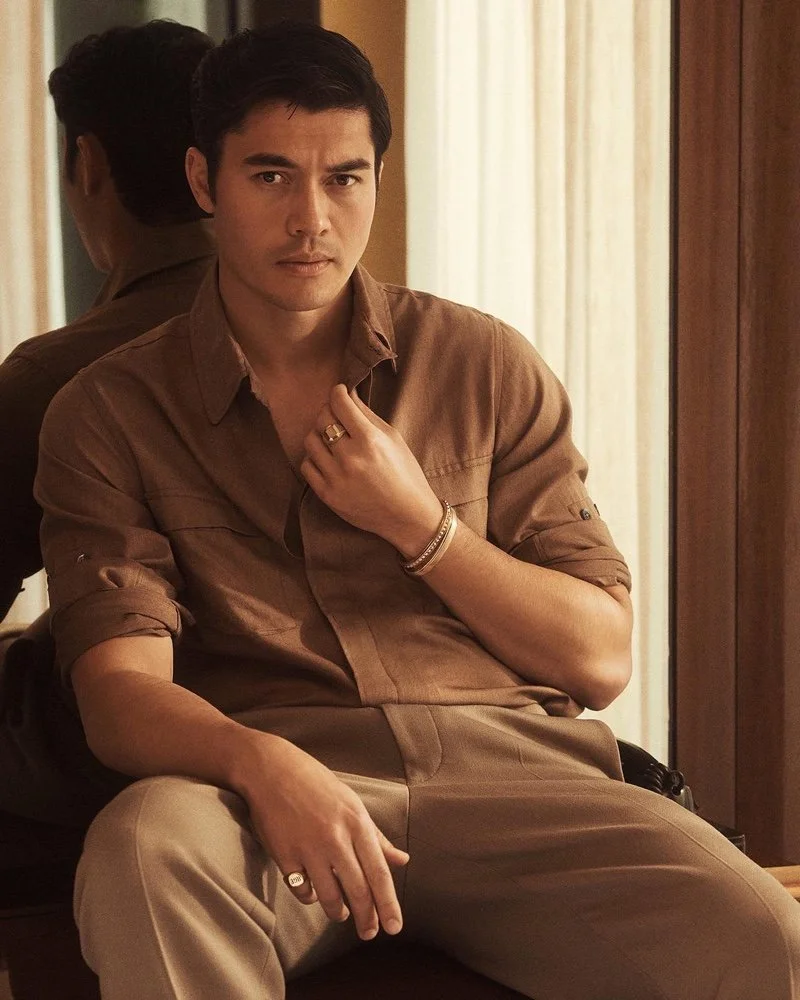 A man with dark hair and light skin sitting in front of a mirror, wearing a brown shirt with rolled-up sleeves and beige pants, with a serious expression and jewelry on his fingers, wrist, and neck.
