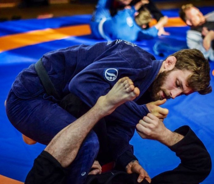 Two men practicing Brazilian Jiu-Jitsu on blue mats, one in a blue gi applying a choke hold on the other in black gi, with a colorful background of other practitioners.