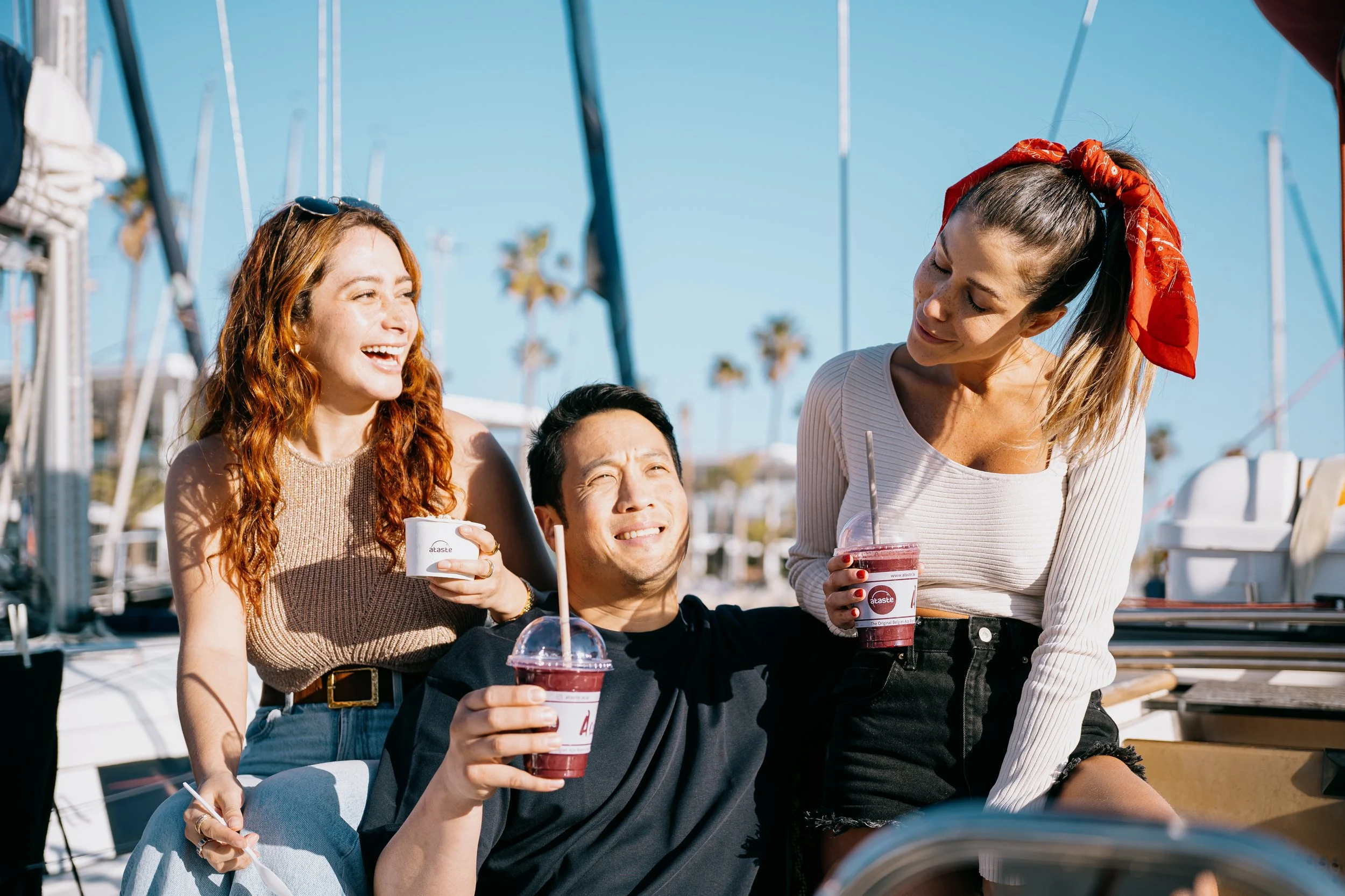 Three friends enjoying drinks and talking at a marina with sailboats and palm trees in the background on a sunny day.
