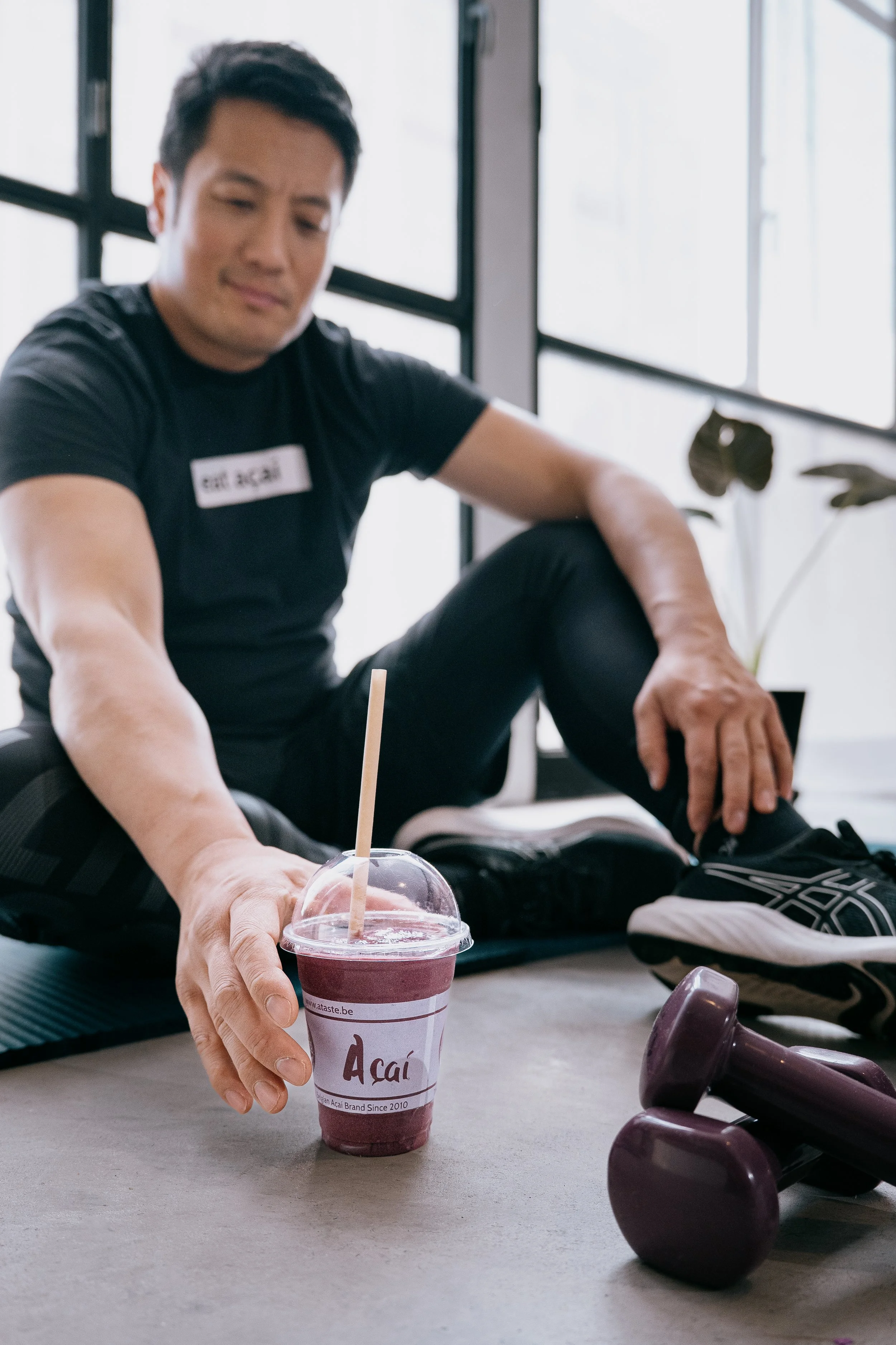 A man sitting on a yoga mat in a bright room, reaching for a cup of acai smoothie, with dumbbells and a plant nearby.