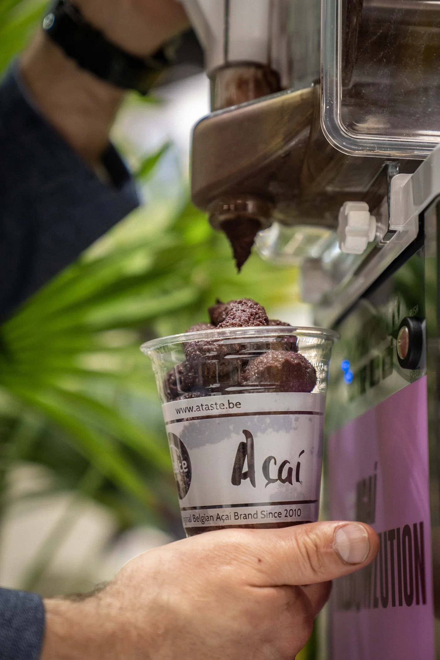 Close-up of a hand holding a clear plastic cup filled with purple acai berry sorbet. The cup has a white label reading 'www.ataste.be' and mentions it is a Belgian acai brand since 2010. A person is operating an ice shaver machine in the background.