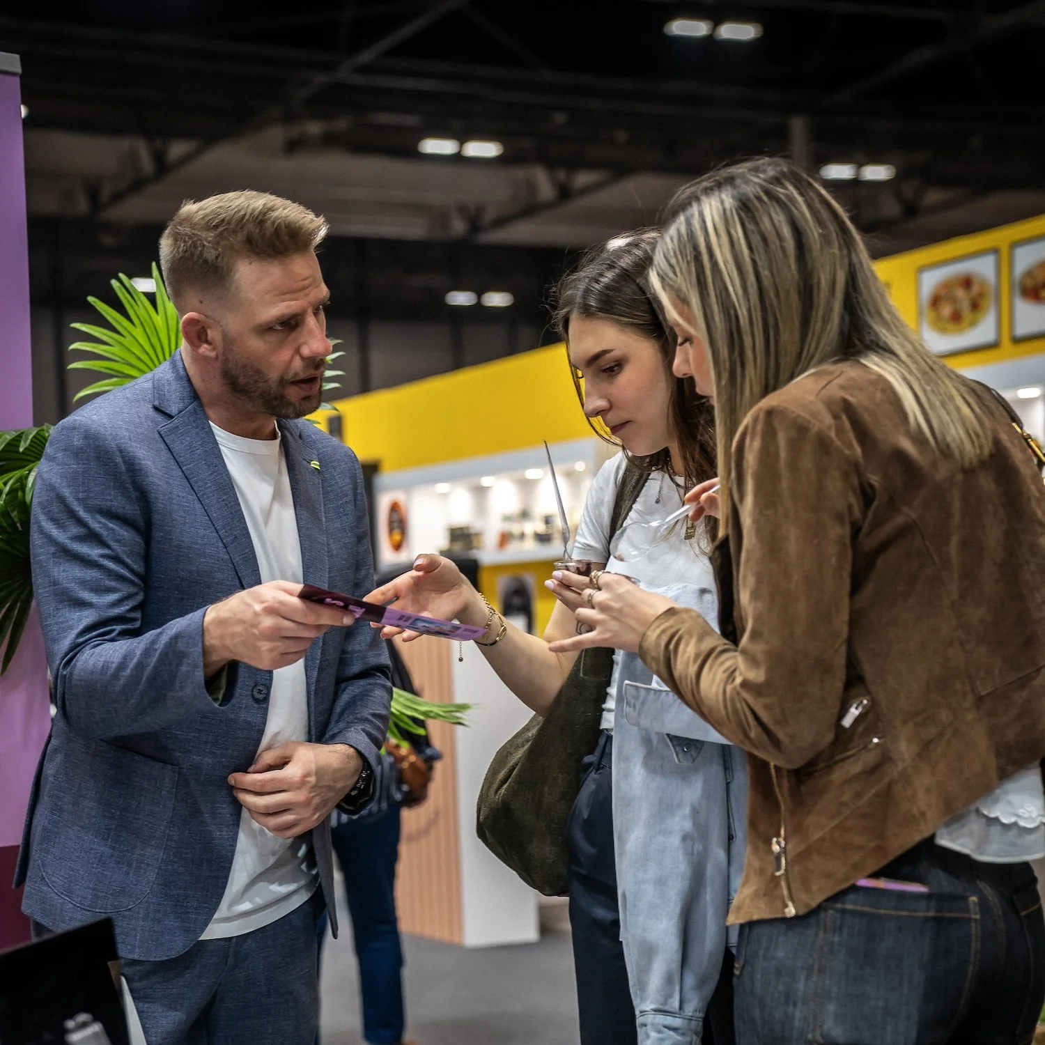 Three people engaged in conversation at an indoor event or exhibition, two women and one man, with a yellow and purple background and a potted plant.