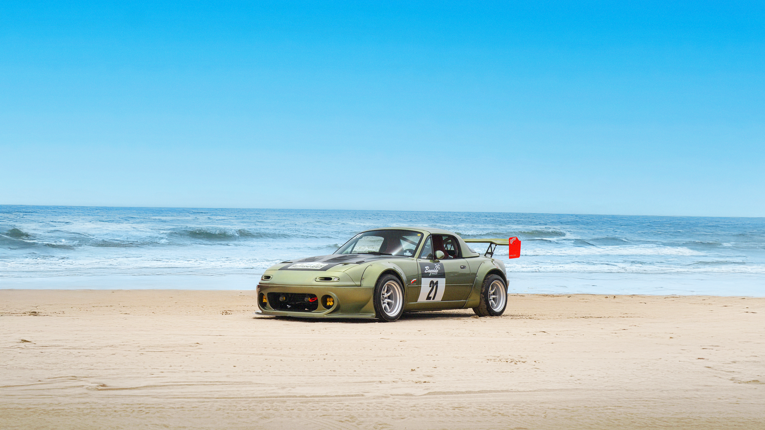 A Mazda Miata racecar sits on the beach as waves crash in the background on a crystal clear day. Part of the Bayside California cruise up the west coast, as featured in Bayside Magazine published by Carrara Media.