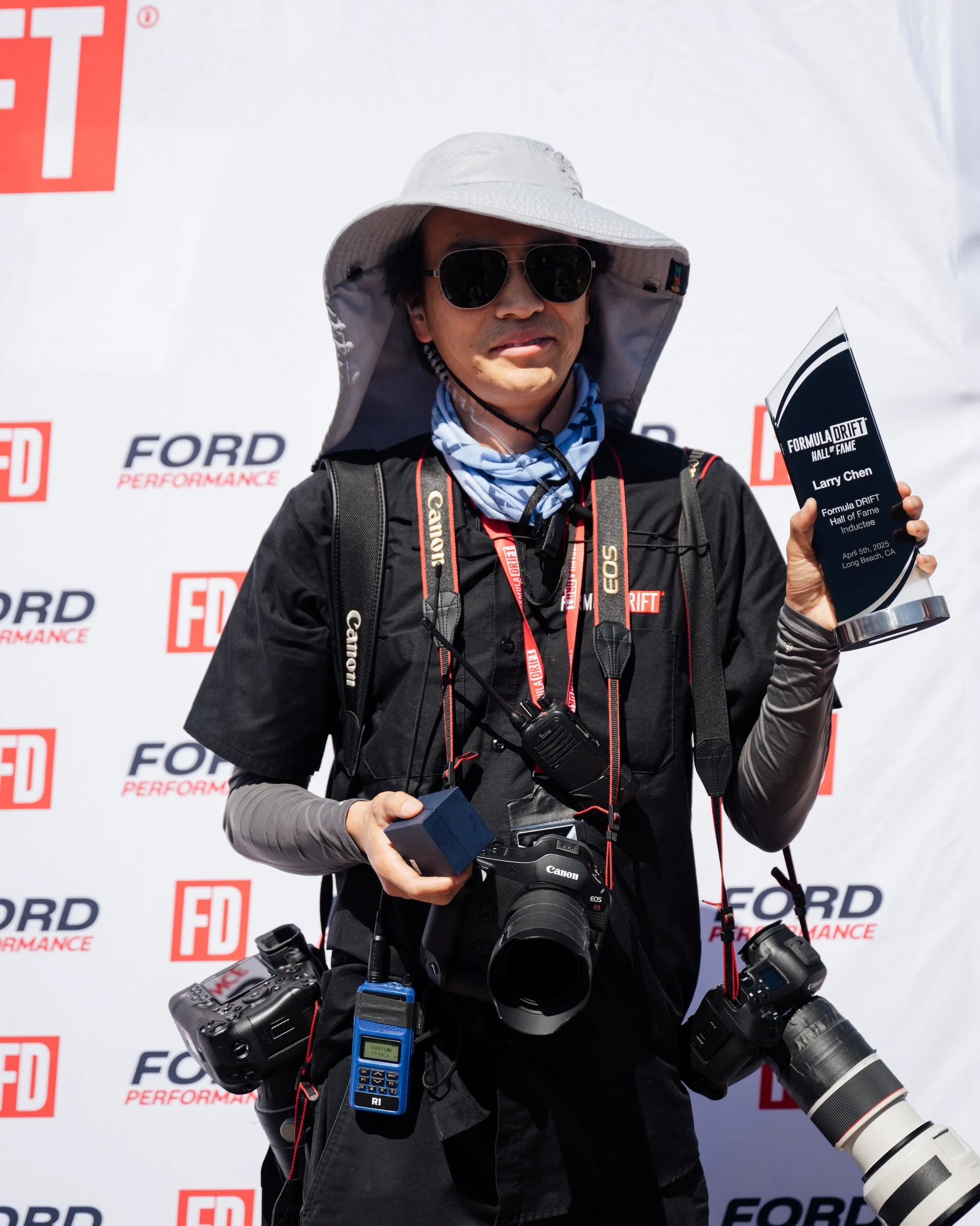 Larry Chen, author of "Life At Shutter Speed," pictured holding his Formula Drift Hall of Fame trophy while draped in multiple cameras around his neck. Published by Carrara Media, 2025.