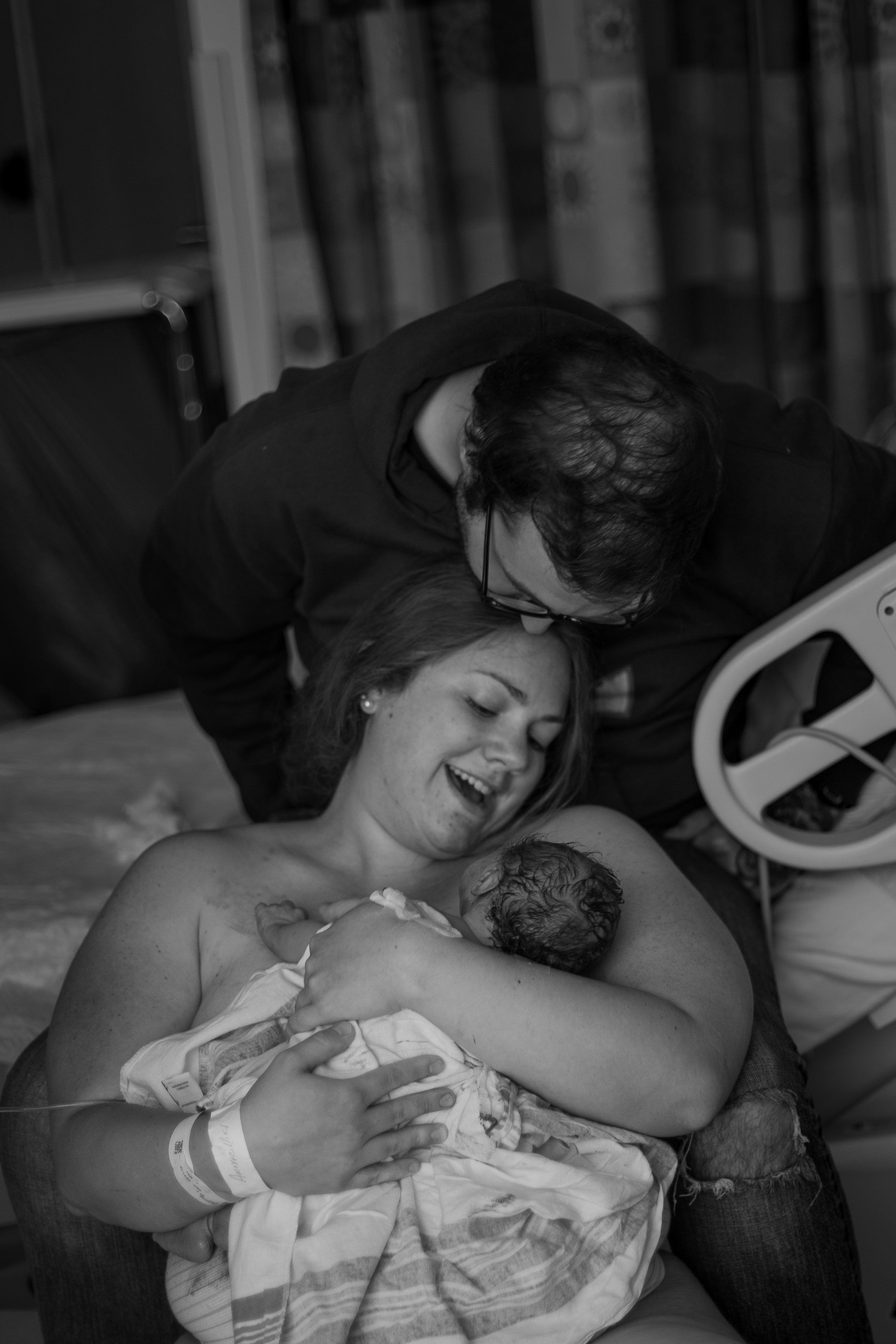 Newborn baby held by mother who is being kissed on the forehead.