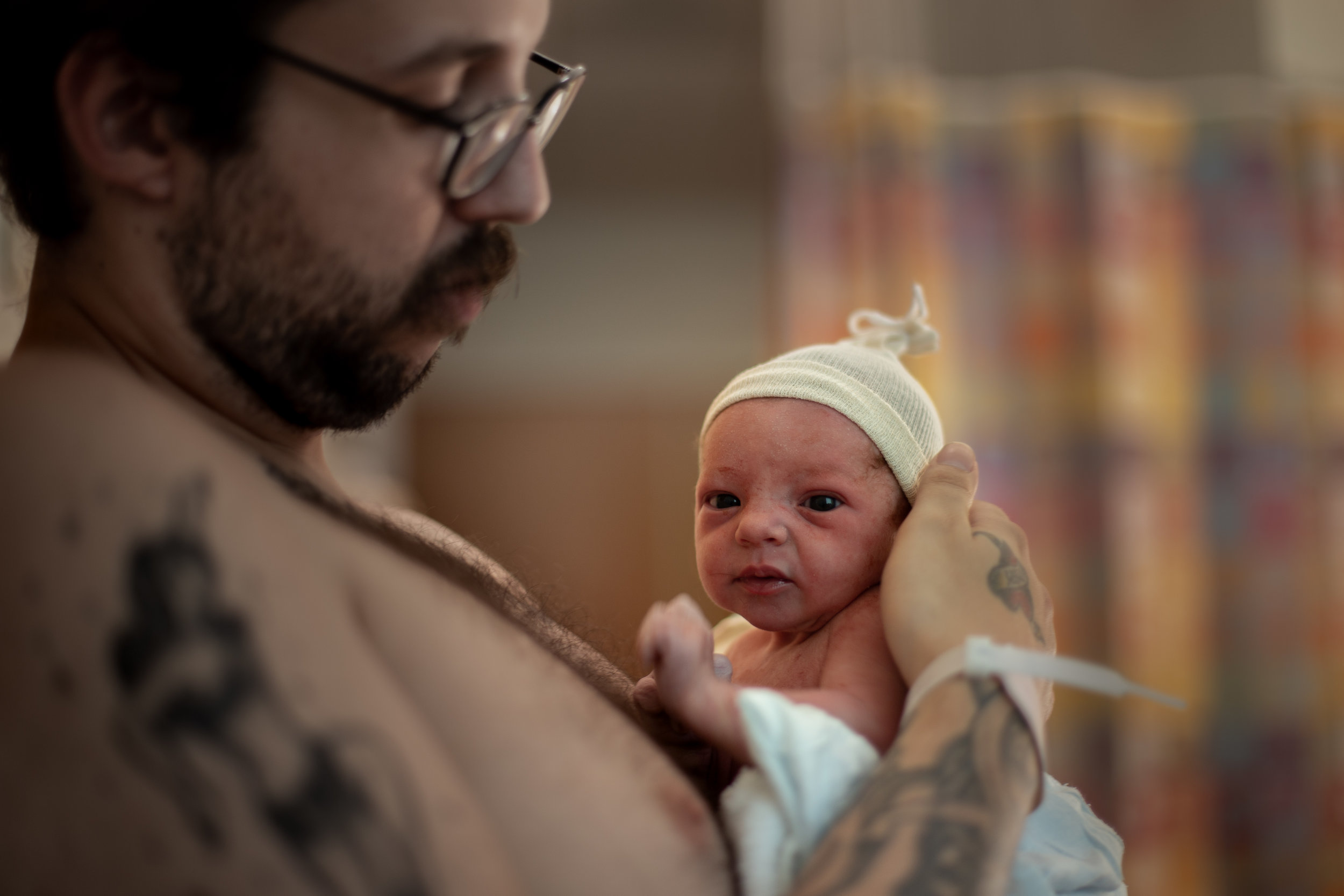 Dad with tattoo holds a newborn who is holding his head up and looking into the camera.