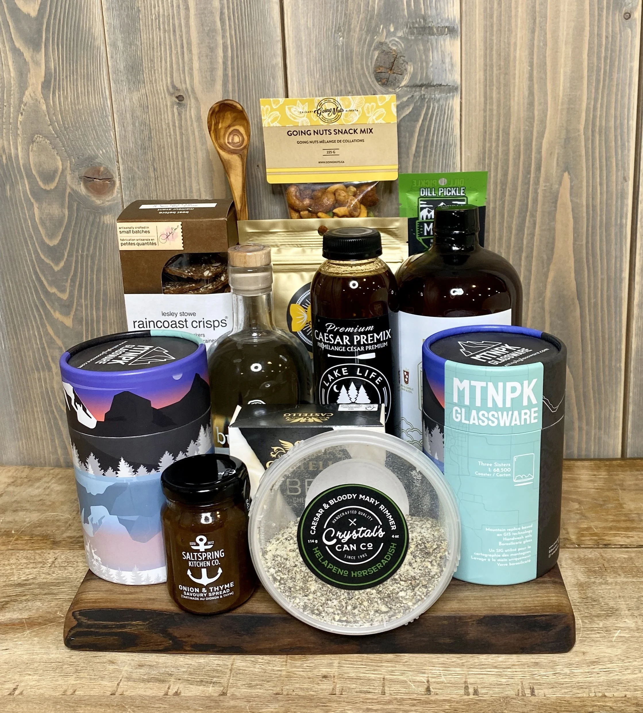 Assortment of gourmet food and drink items including crisps, oil, snack mix, drink mixers, condiments, and specialty glassware on a wooden surface.