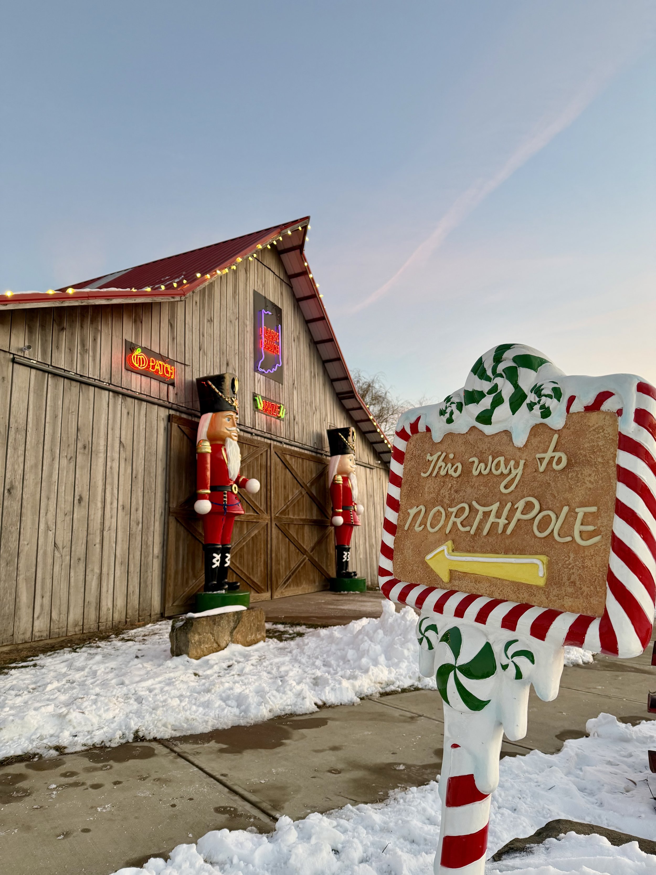 Decorative sign with candy cane border and arrow pointing to the north pole, in front of a wooden building with holiday decorations and nutcracker statues outside, snow on the ground.