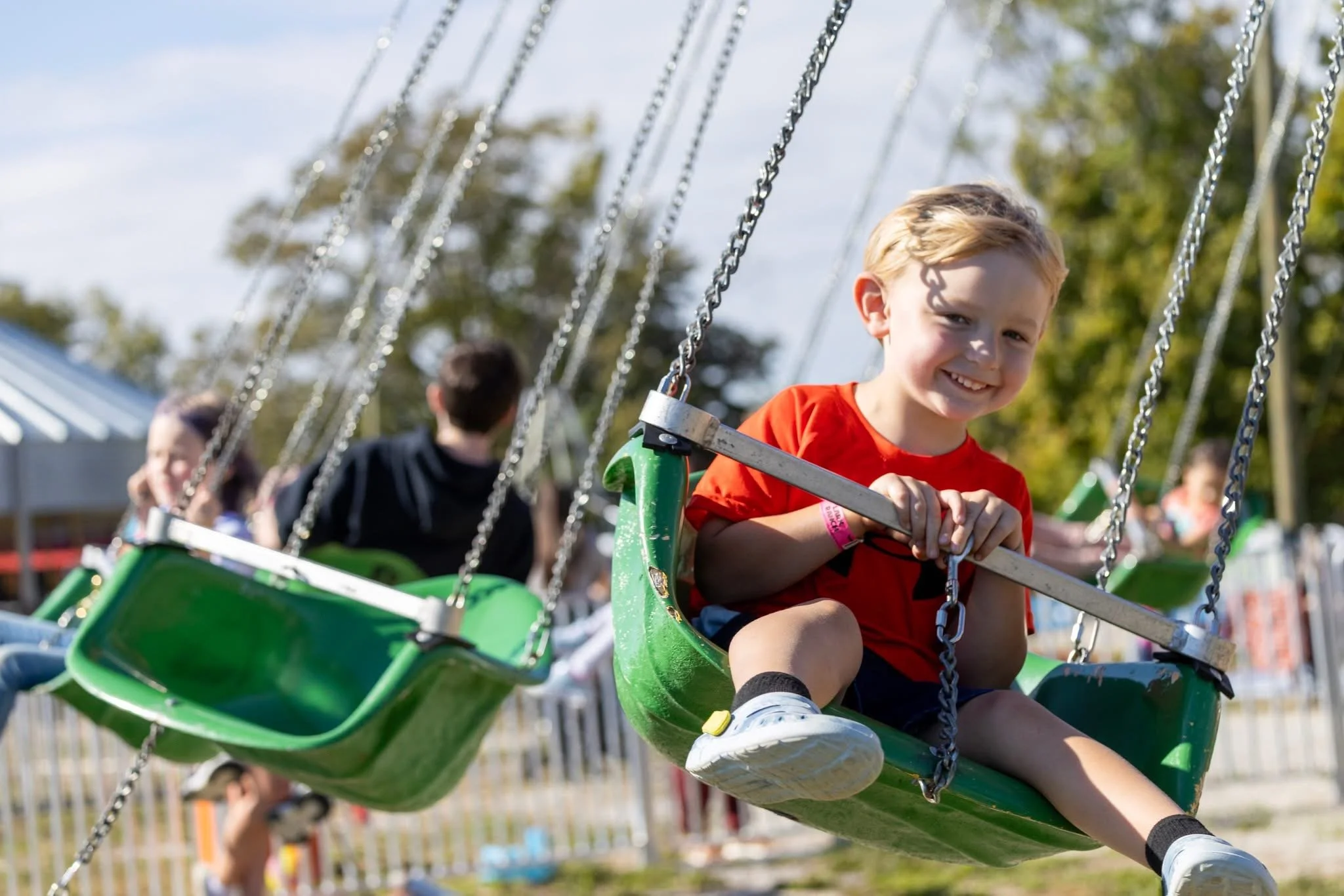 A young boy with blonde hair and a red shirt smiling while riding a green swing ride at an amusement park, with other children in the background.