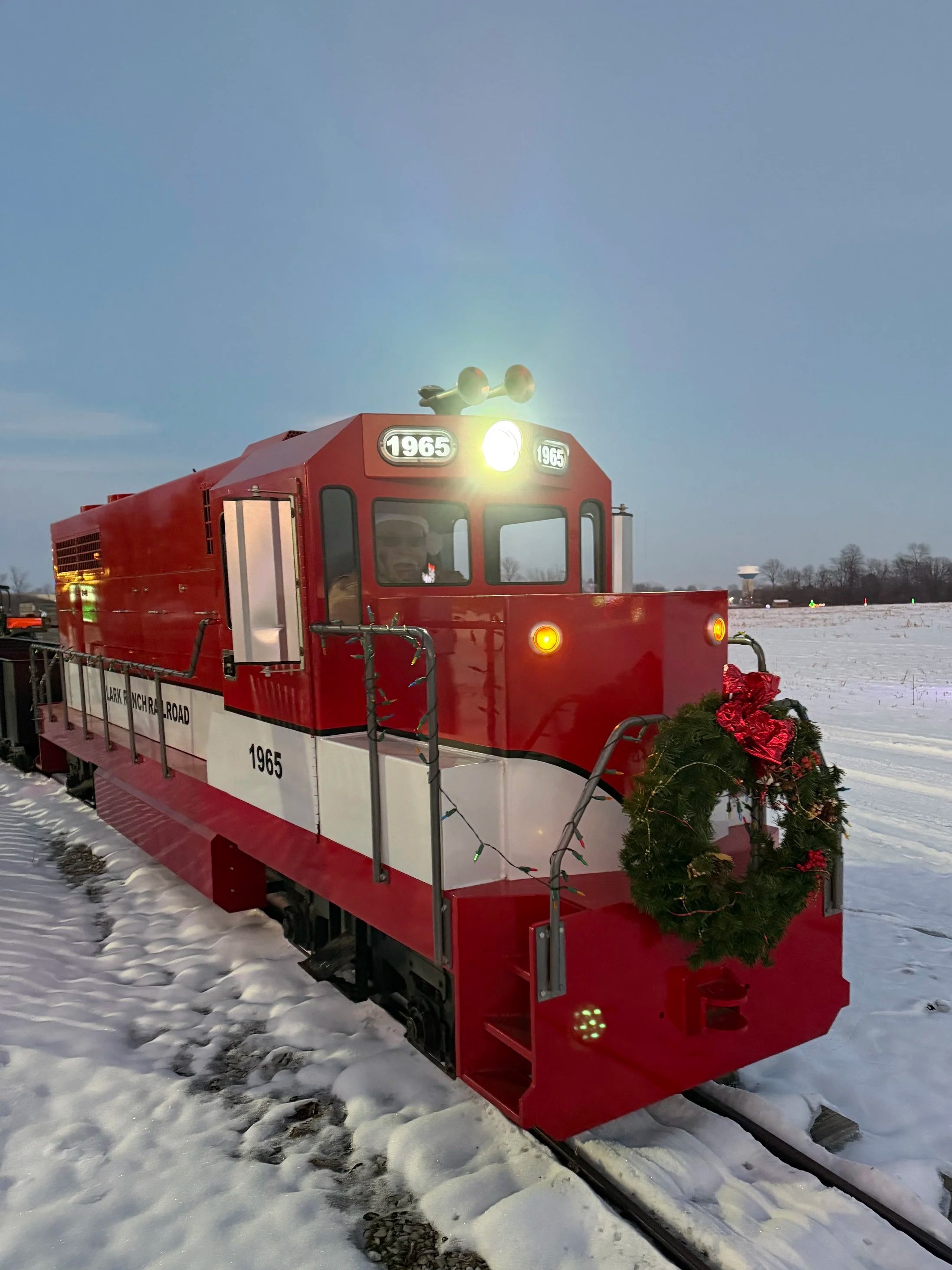 A red trolley decorated with a Christmas wreath at the front, situated on snow-covered tracks outdoors in a wintry landscape.
