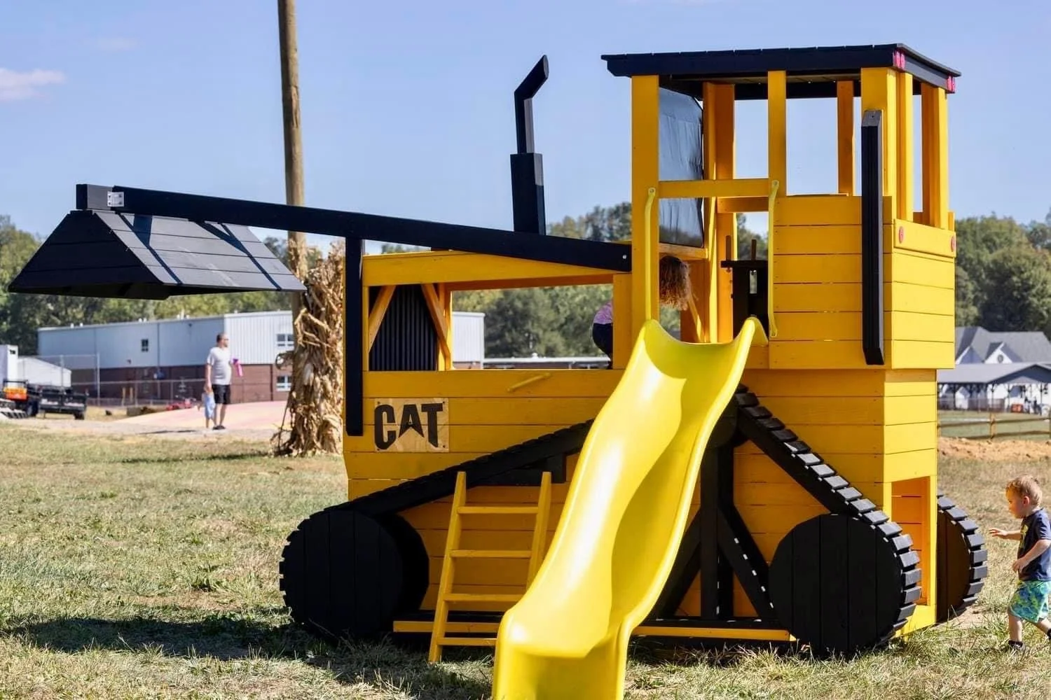 A playground structure designed like a yellow train with black wheels, a yellow slide, and a small staircase, with children playing around it outdoors on a grassy area.