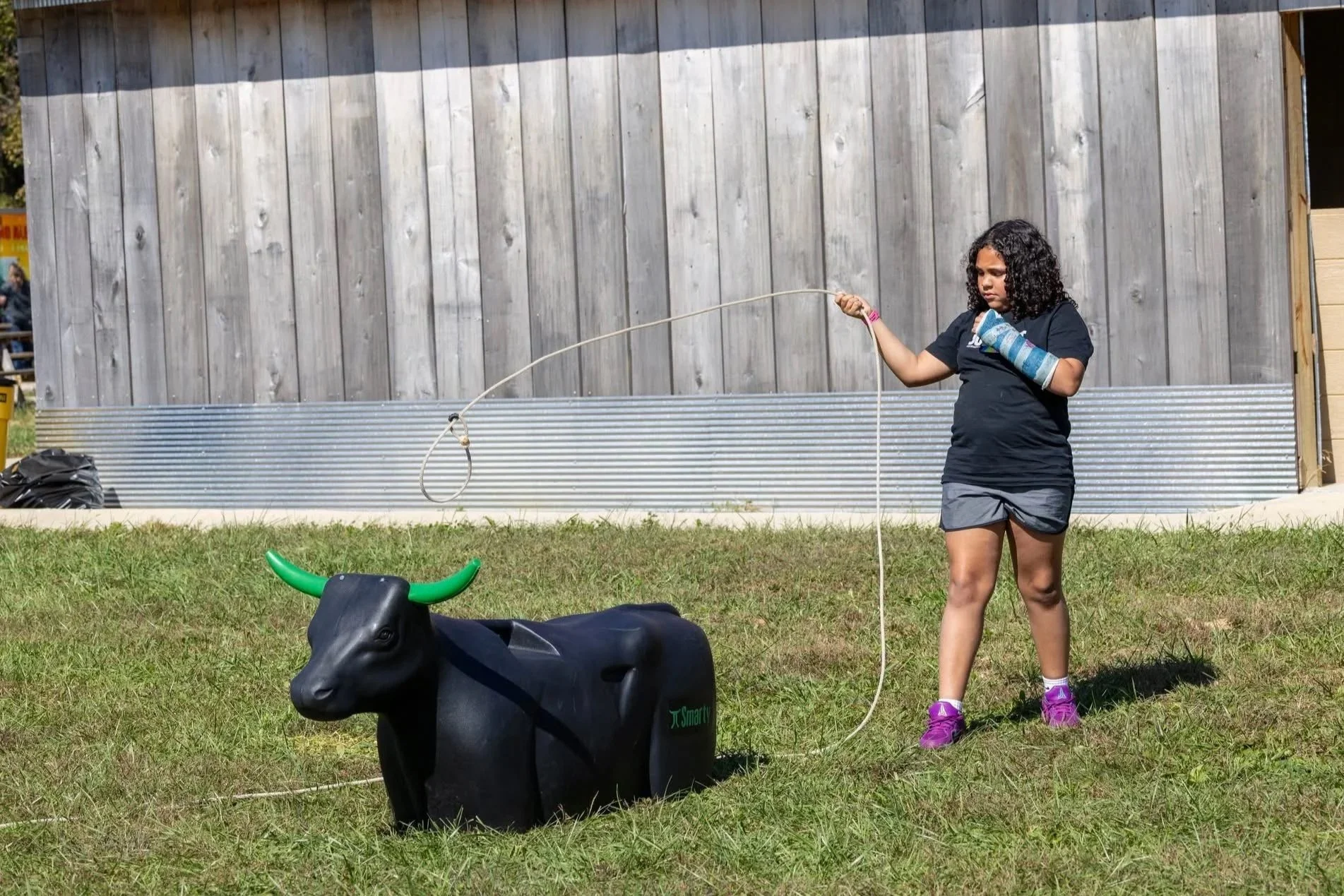 A young girl with curly hair, wearing a black t-shirt, gray shorts, purple shoes, and a blue arm cast, holding a long leash attached to a black robotic cow figure on a grassy area outside, with a wooden building in the background.